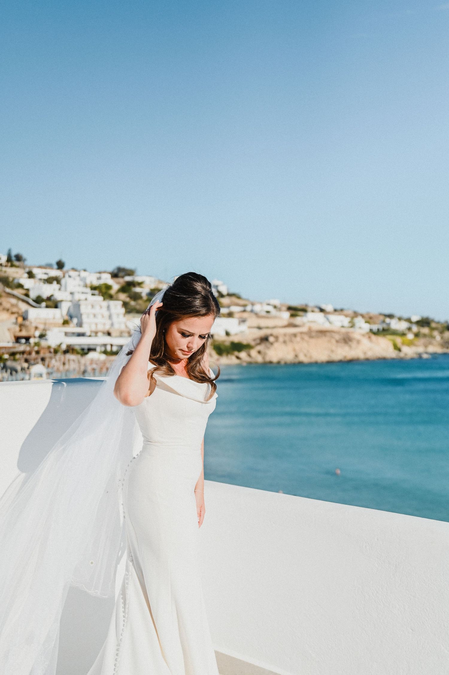Elegant bride in fitted white gown stands against blue Mediterranean Sea backdrop.