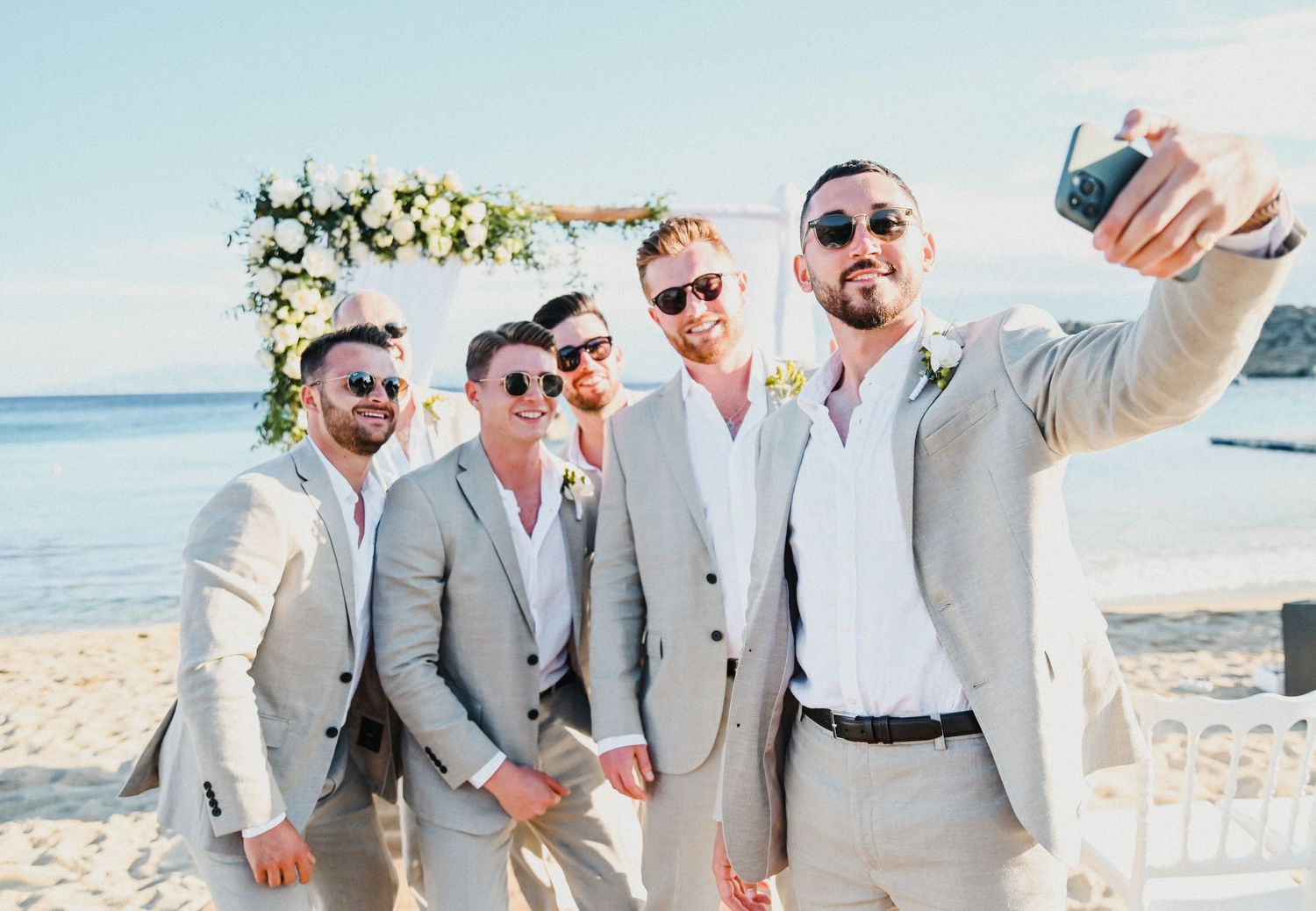 Groomsmen in light gray suits pose for a selfie on a sunny beach with floral arch backdrop.