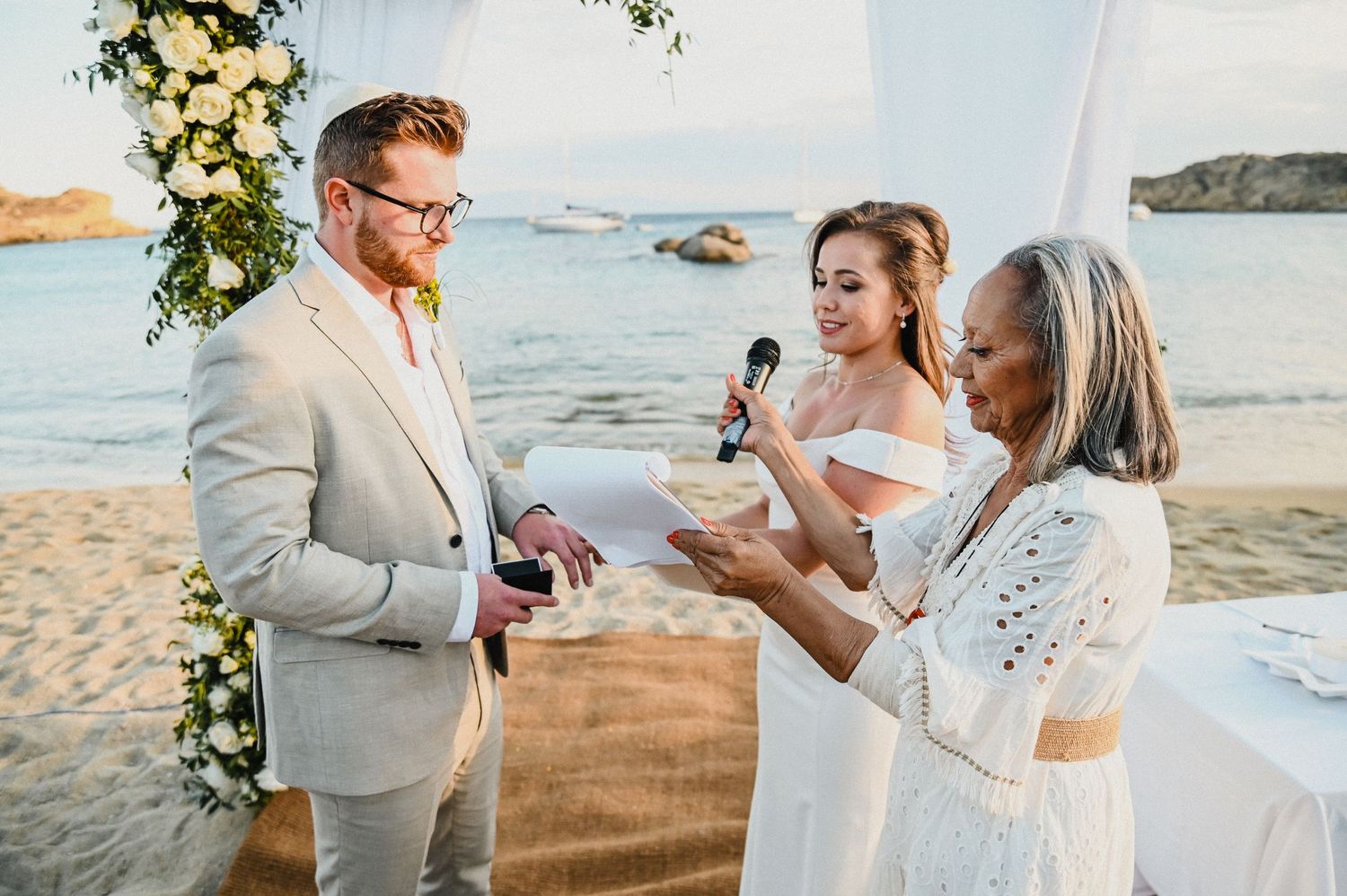 Joyful wedding ceremony moment as couple shares laughter with officiant under floral arch by the ocean.