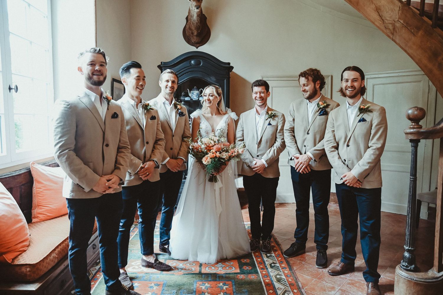 Wedding party poses together in neutral tones by a curved staircase and mounted deer head decoration.