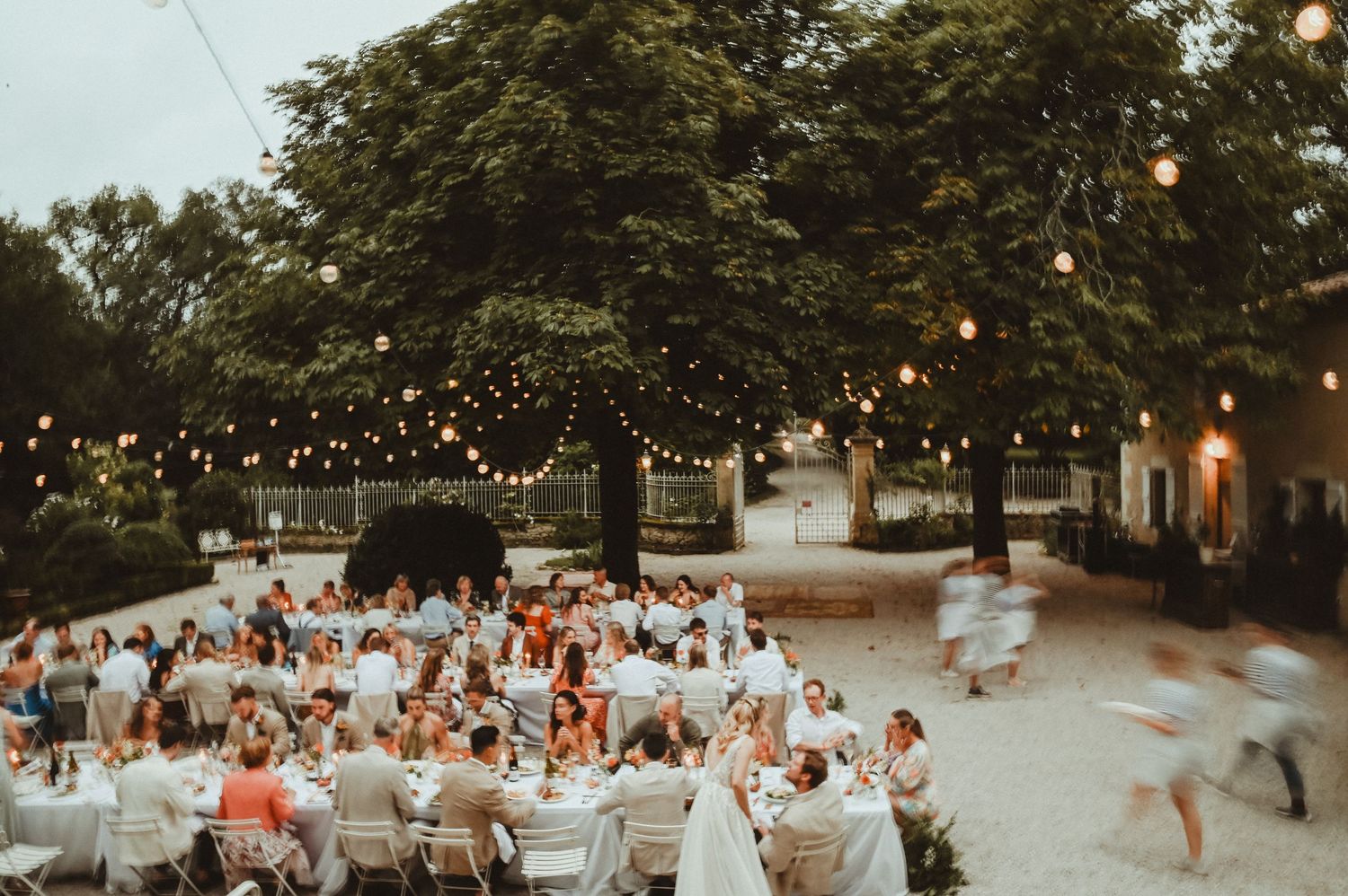 Evening outdoor reception with string lights and white tables under trees at dusk.
