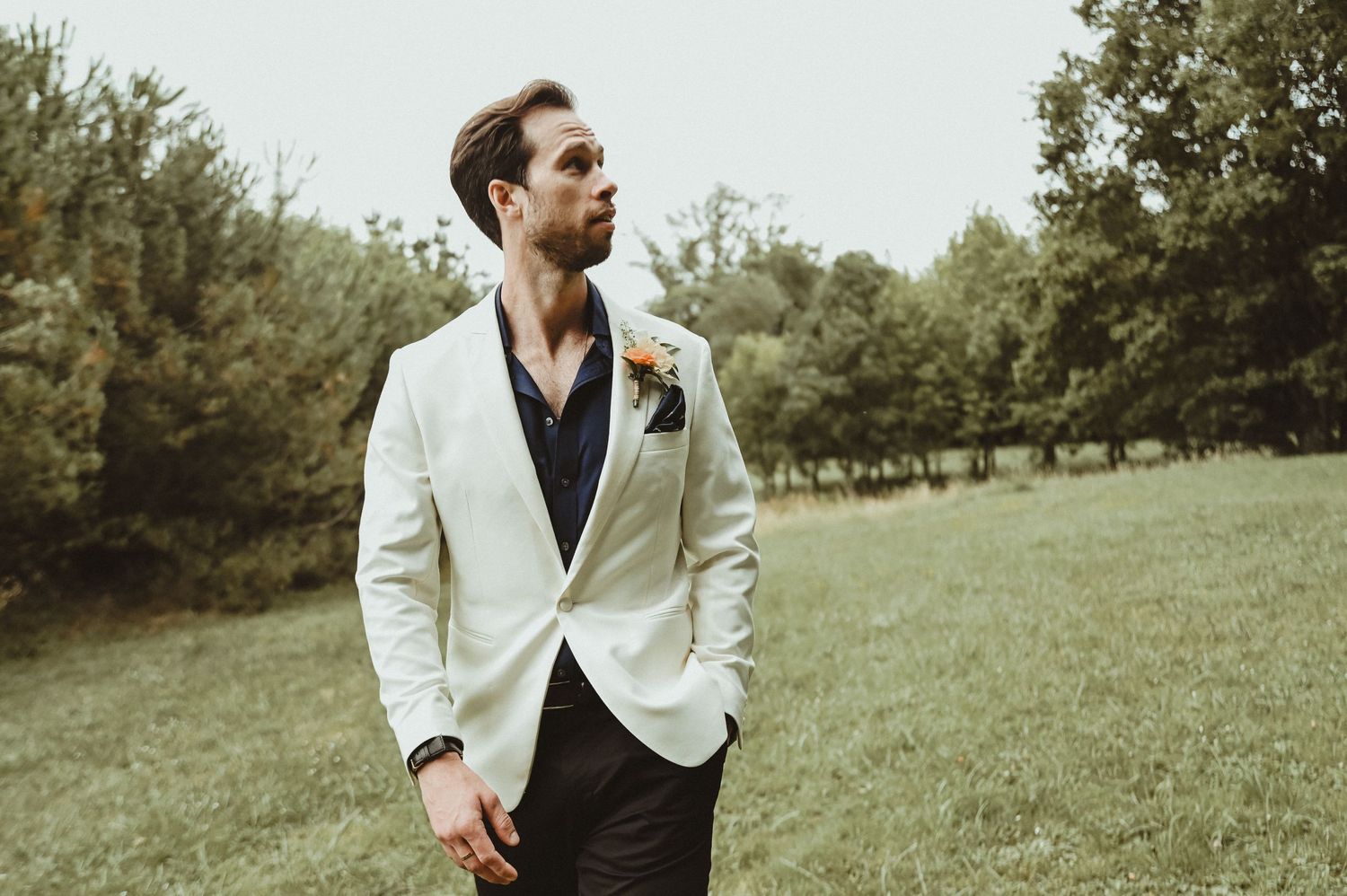 A groom in cream blazer and black shirt poses thoughtfully in an outdoor setting.