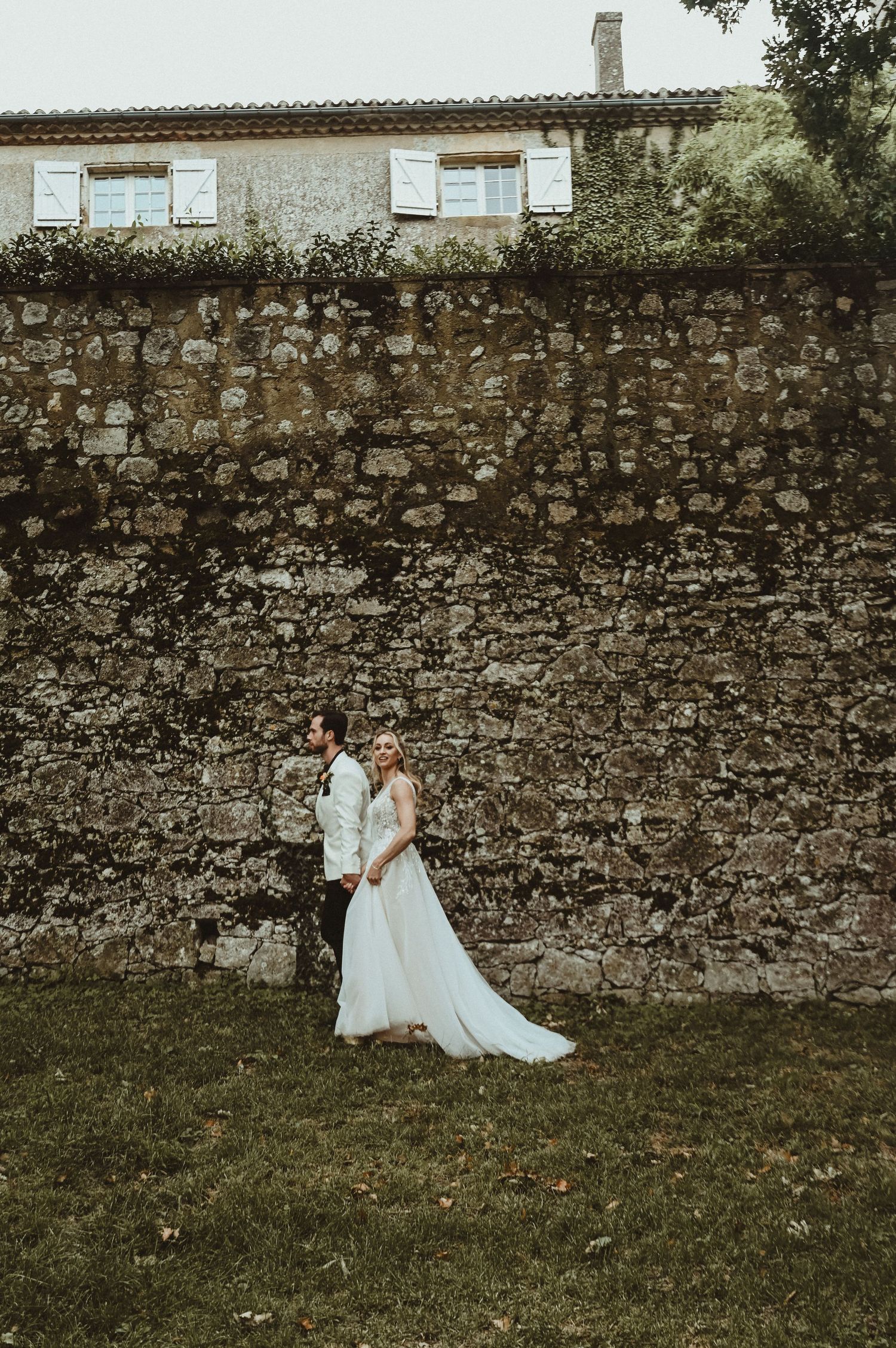 A couple poses against an ancient stone wall outside a French country house.