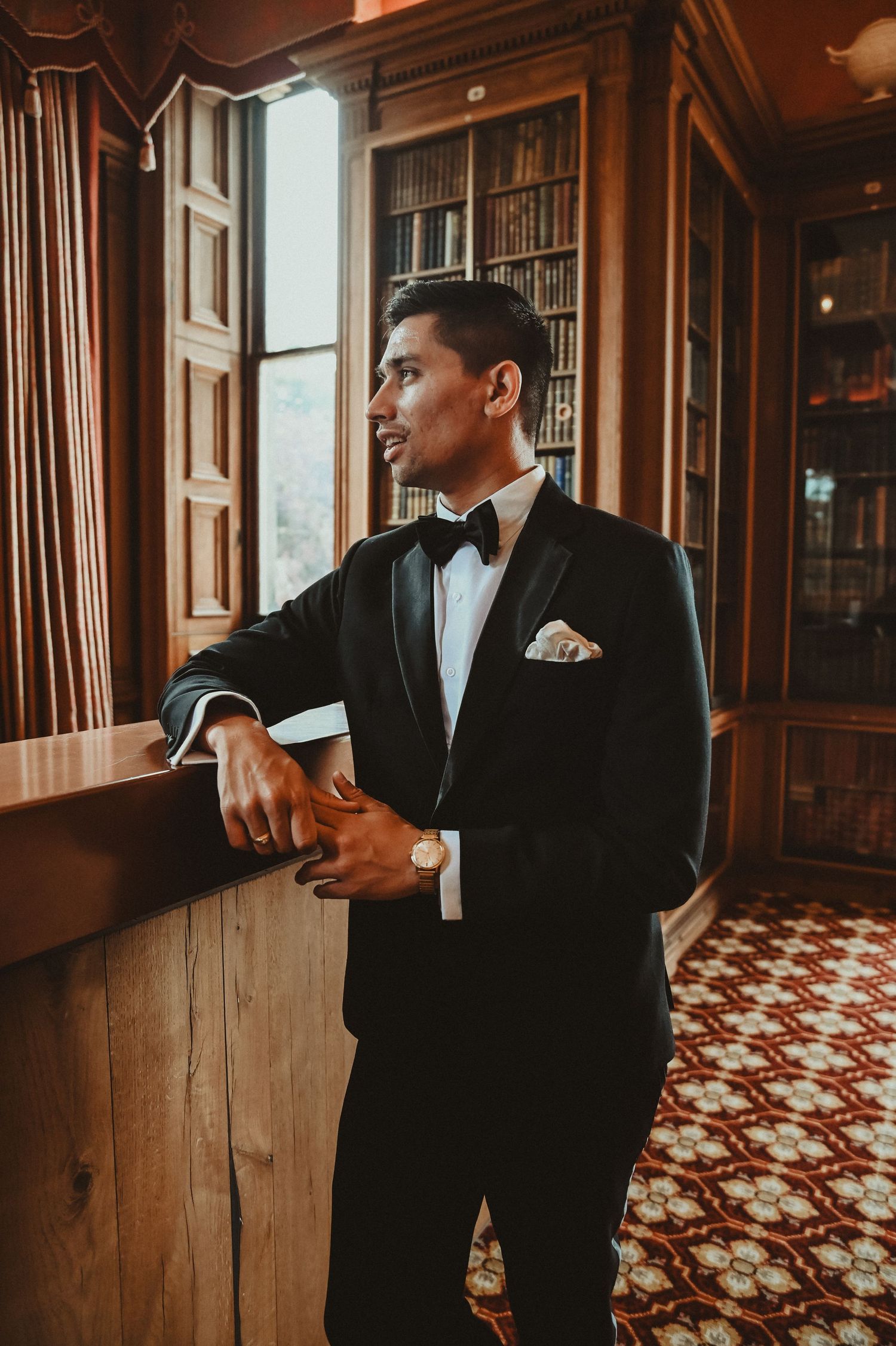 Well-dressed figure in classic black tuxedo and bowtie gazing through window in elegant vintage interior setting.