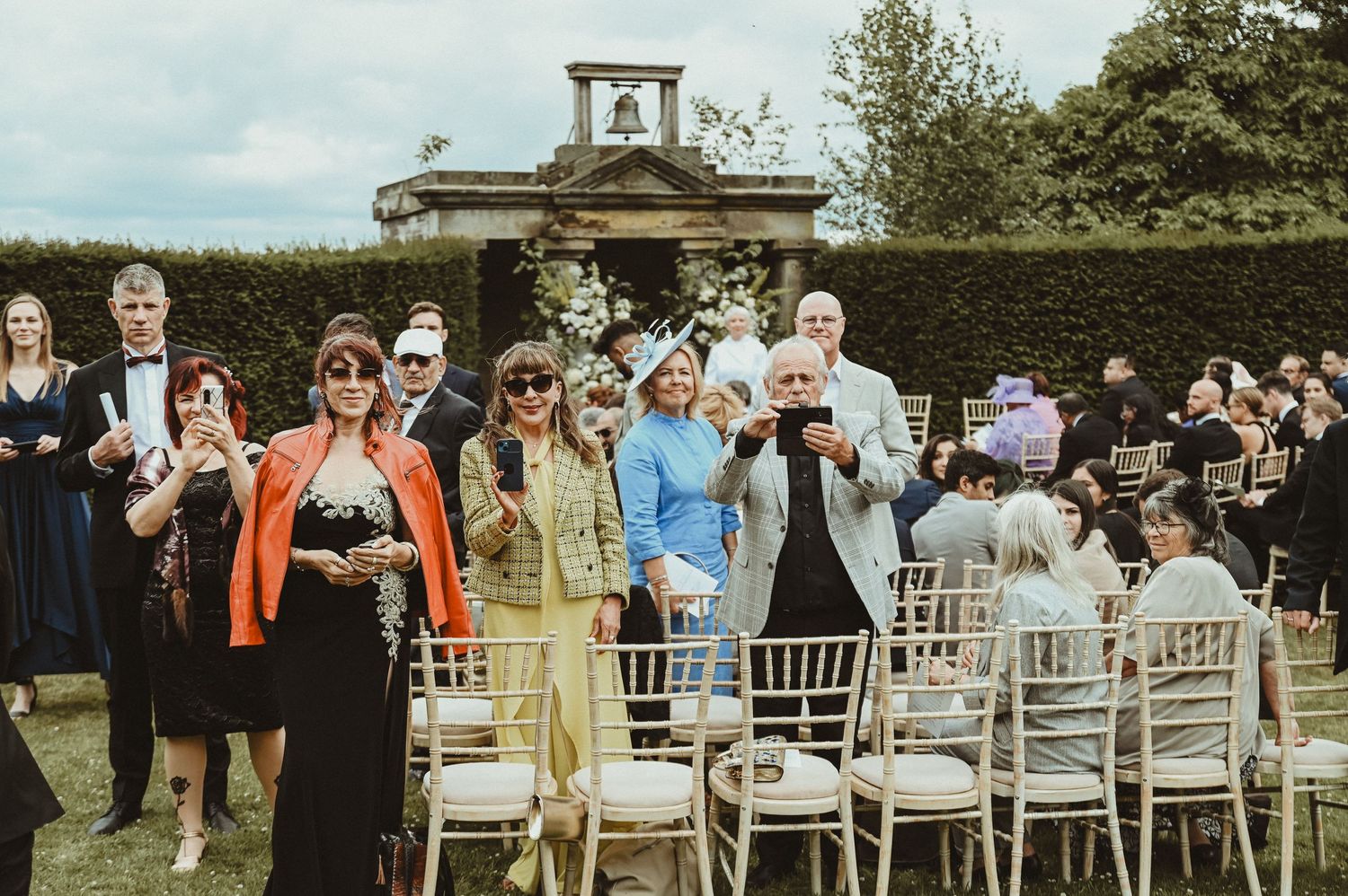 Wedding guests seated in white chairs at an outdoor garden ceremony with ornate architecture.