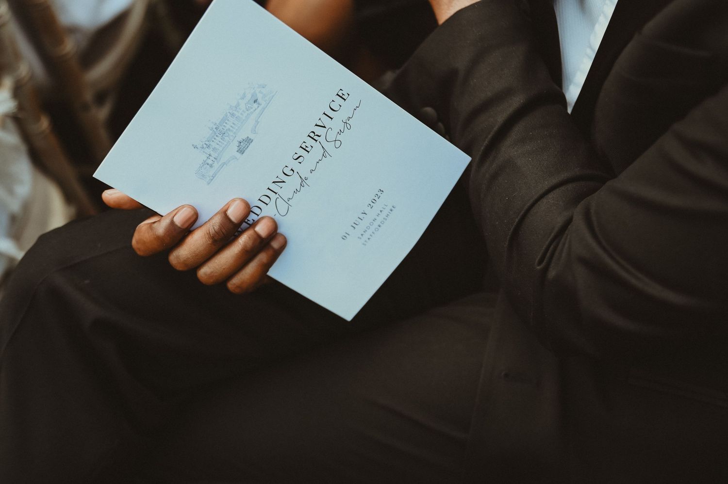 Hand holding a light blue wedding service program against formal attire.