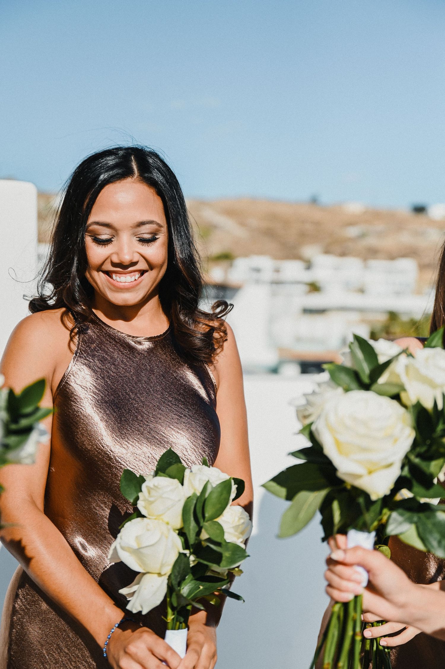 Bridesmaid in a champagne-colored dress holds white roses while smiling outdoors.