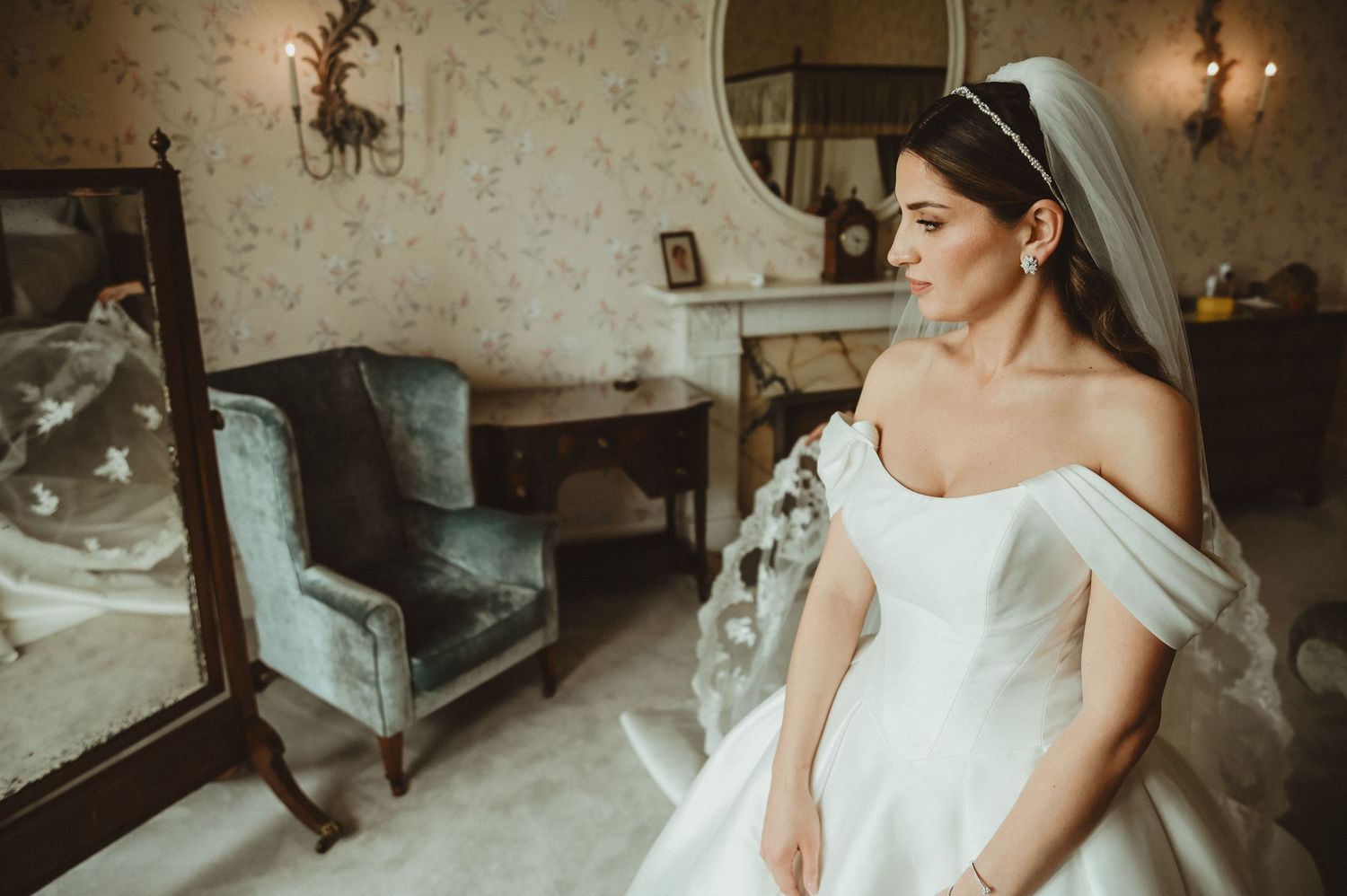 A classic off-shoulder wedding dress photographed in an elegant room with vintage furnishings.