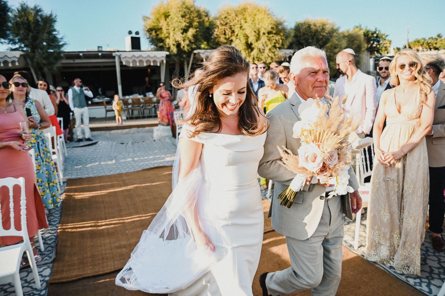 Sun-drenched outdoor wedding ceremony with decorative white chairs and standing guests.
