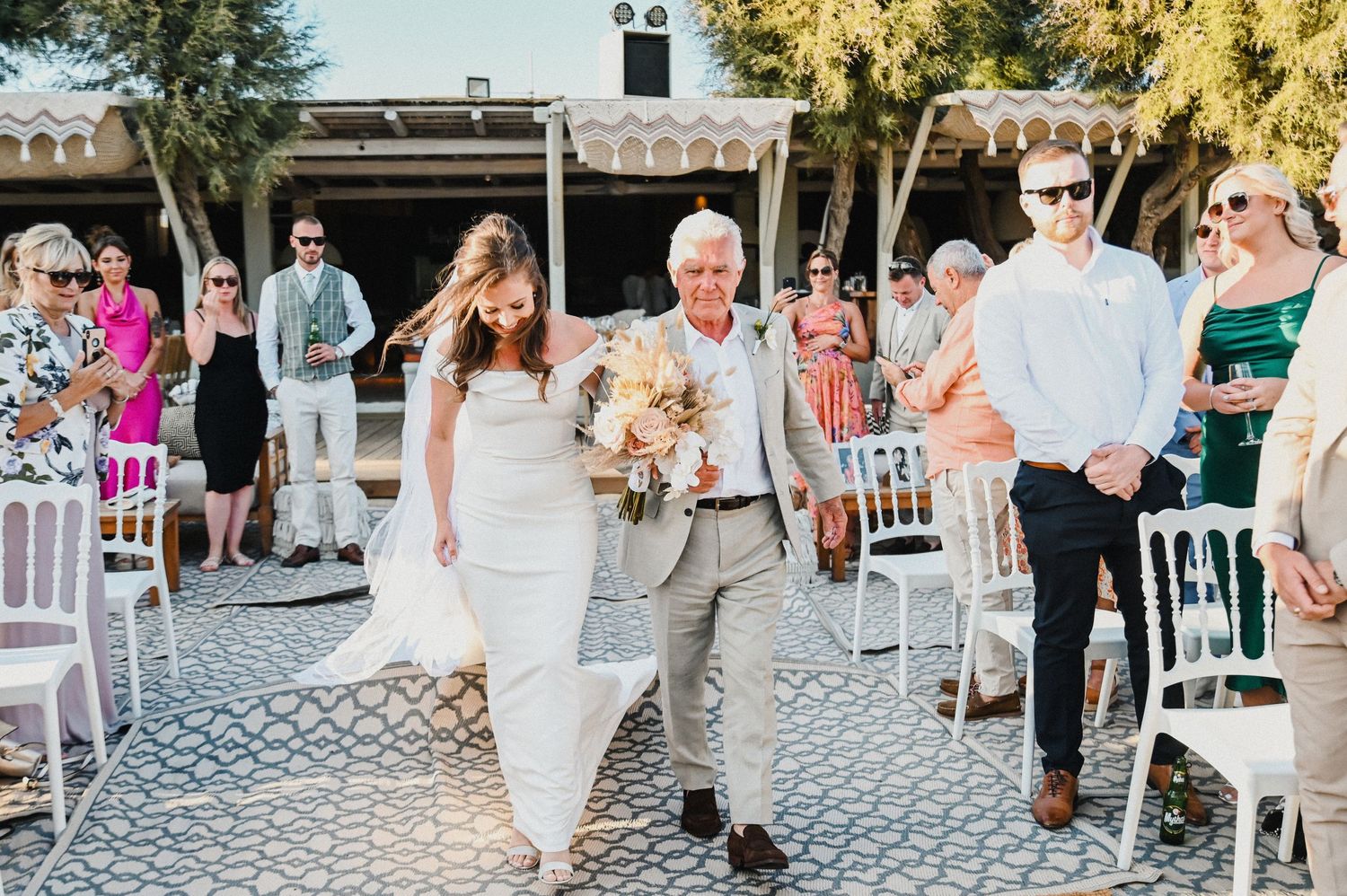 Wedding ceremony procession on cobblestone path with white chairs and standing guests.