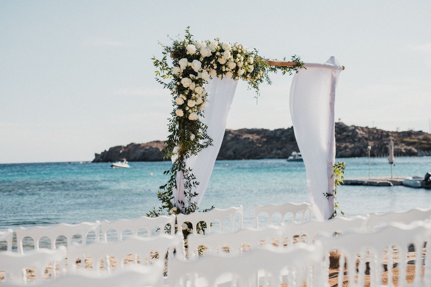 Beachside wedding arch with white fabric and flowers overlooking turquoise Mediterranean waters.