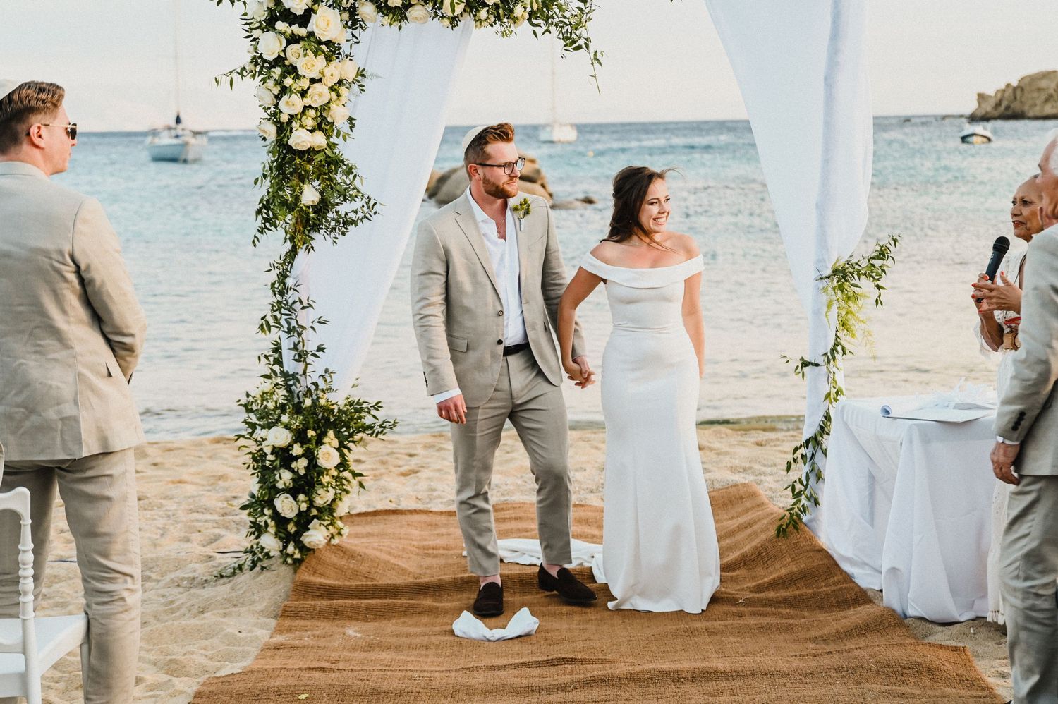 Wedding couple stands together on wooden platform with ocean and floral arch backdrop.