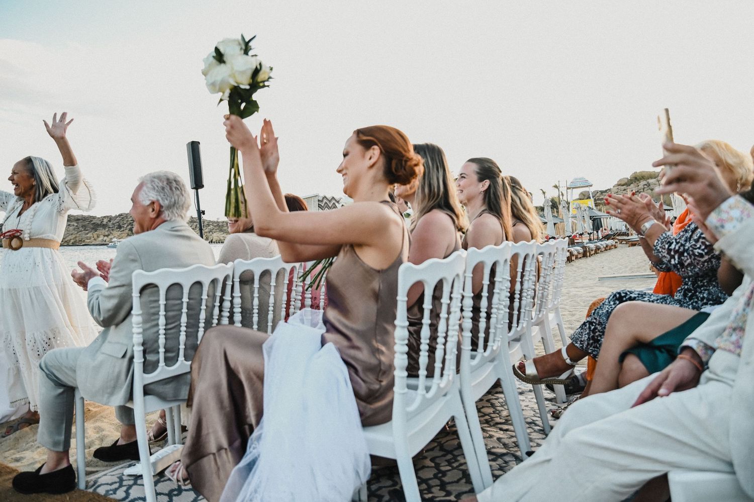 Wedding guests seated in white chairs raise their arms in celebration during beach ceremony.