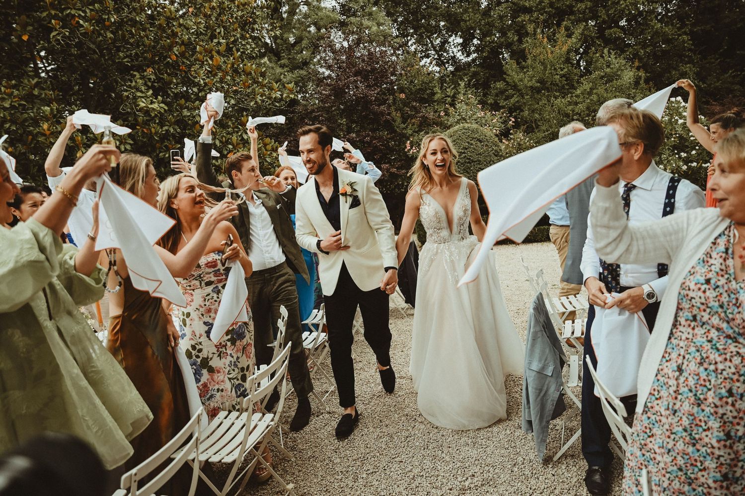Wedding guests wave napkins in celebration as newlyweds walk down a garden path at an outdoor reception.