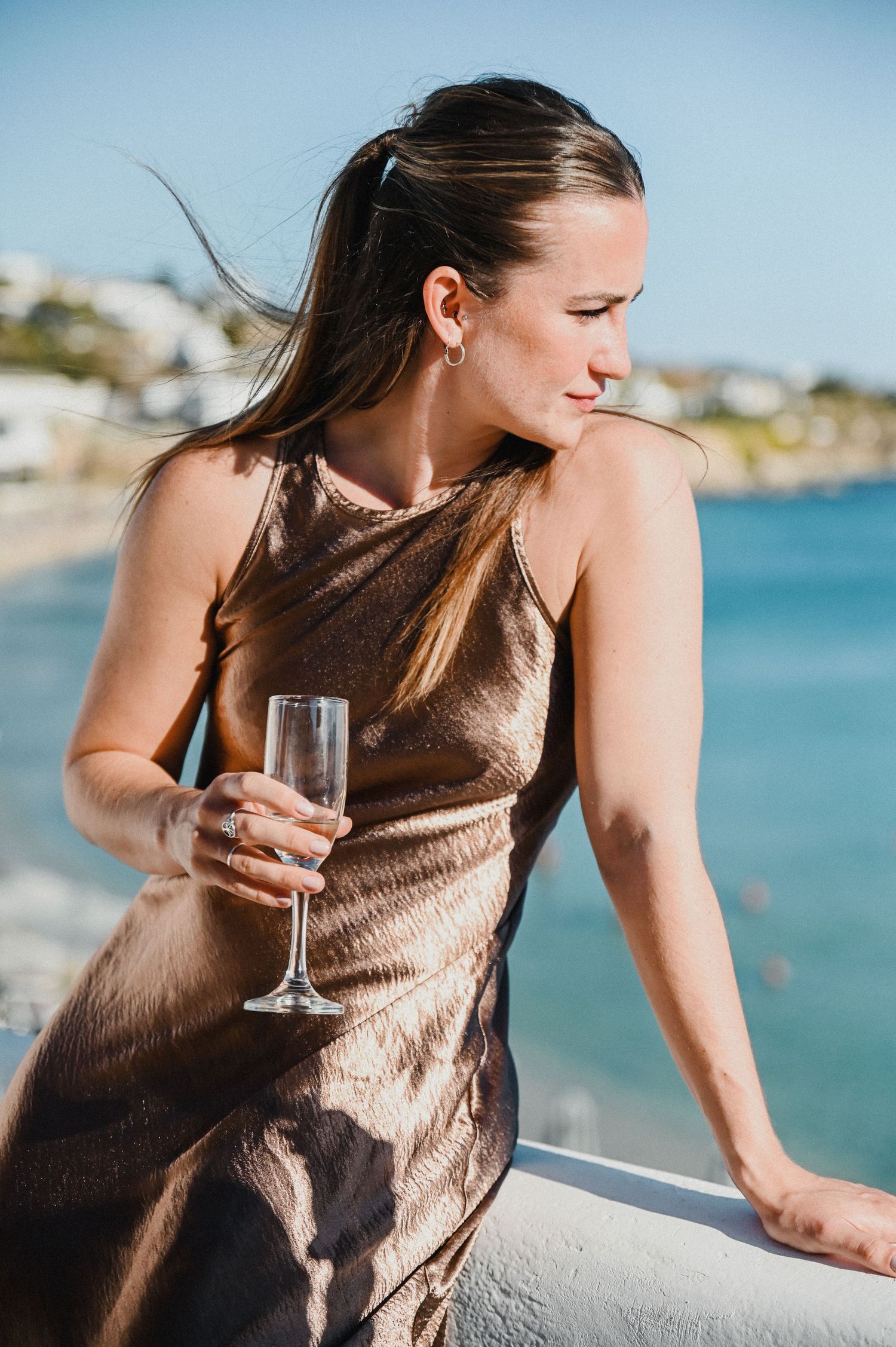 Woman in satin bronze dress holds champagne while enjoying coastal views from a balcony.
