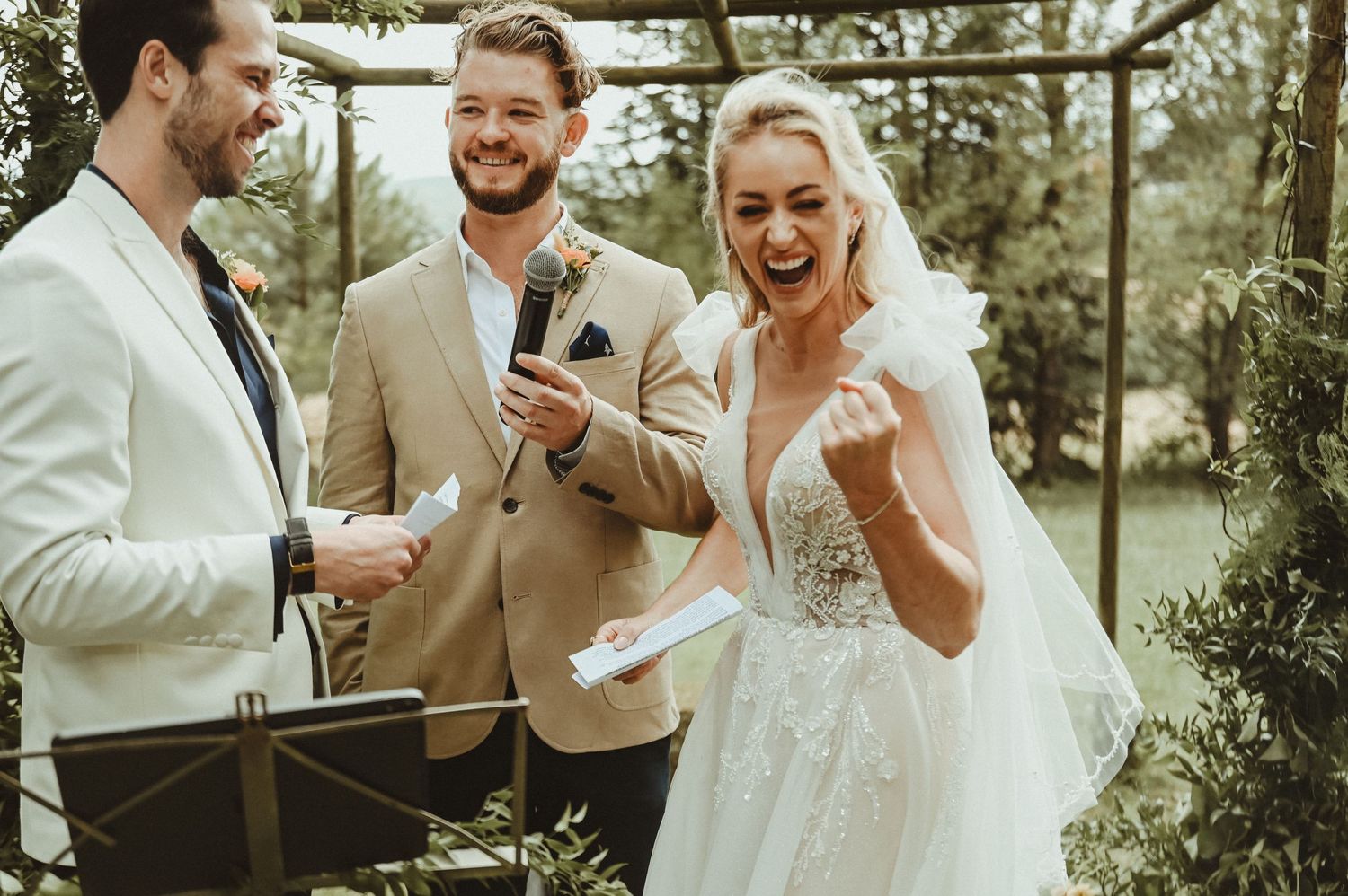 Three wedding party members share a joyful moment during an outdoor ceremony.
