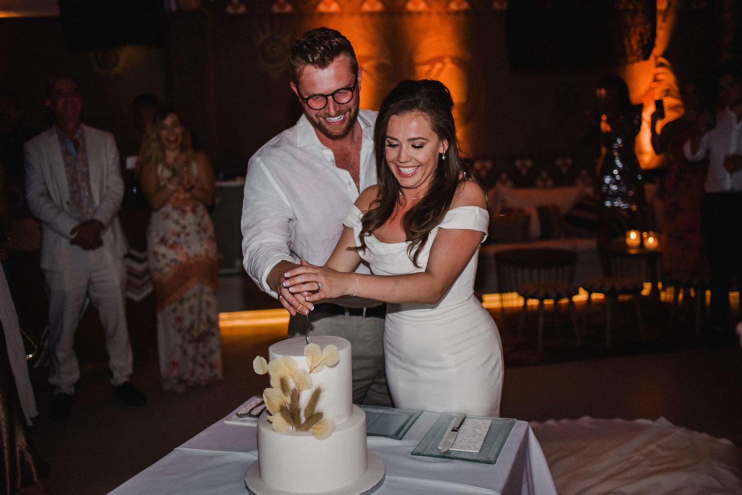 Newlyweds cutting their elegant white wedding cake together in a warmly lit reception venue.