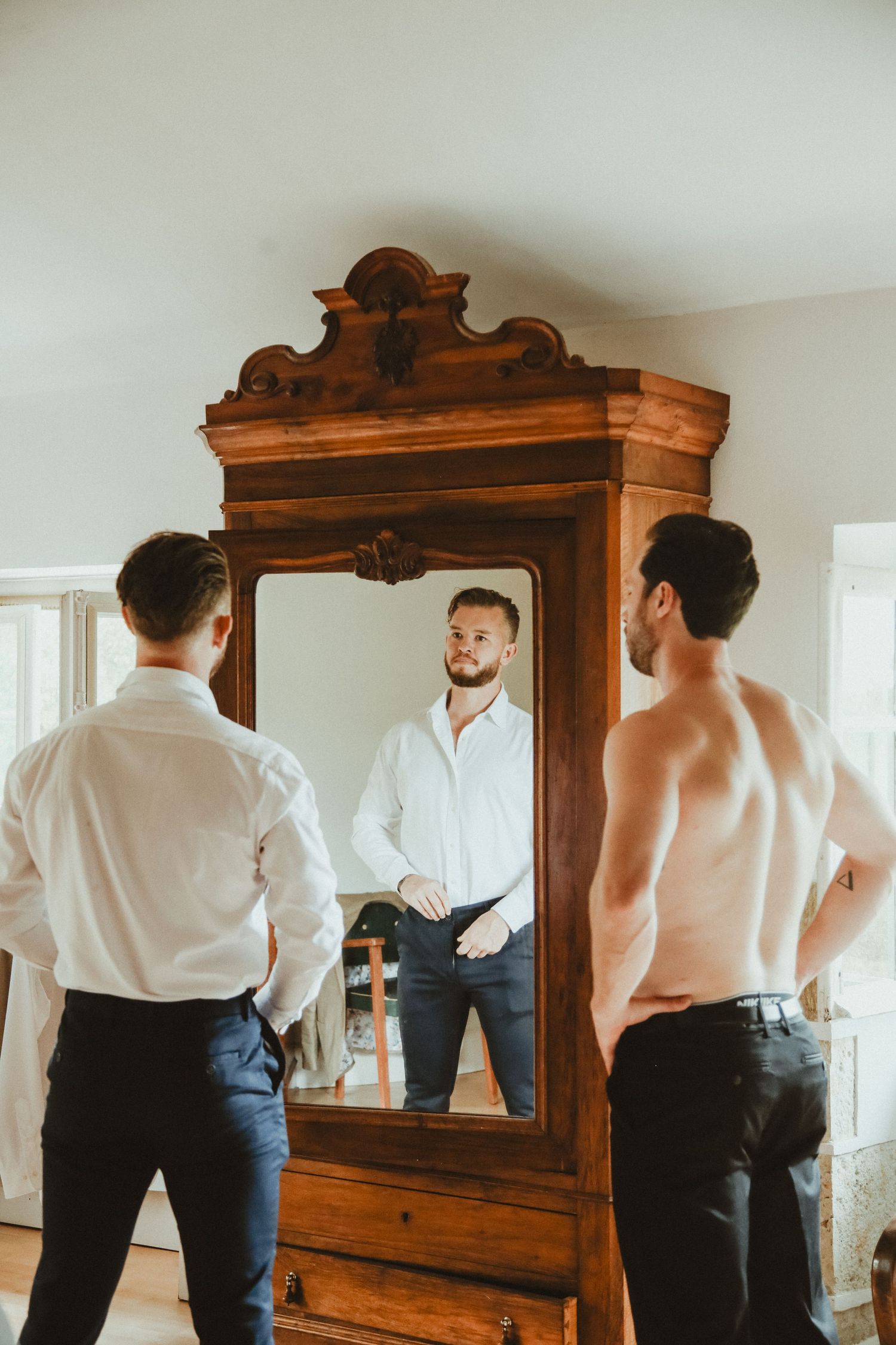 People getting dressed reflect in an ornate vintage wooden armoire mirror.