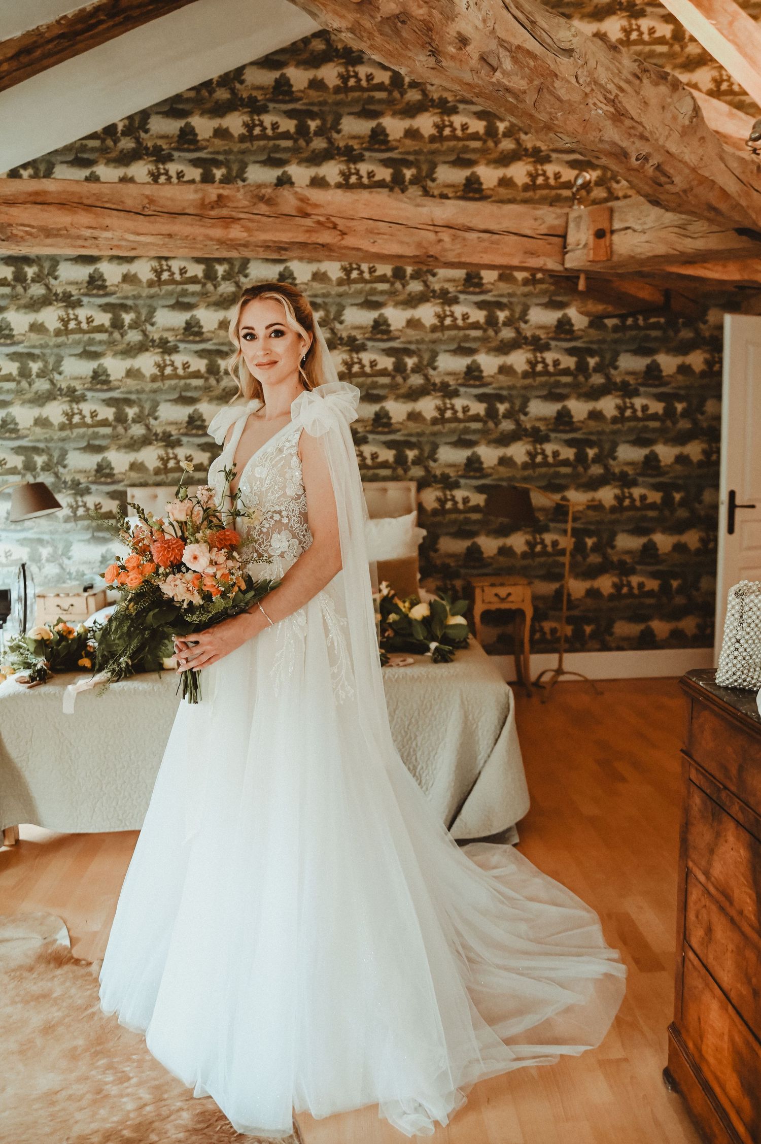 Full-length portrait in a rustic room with wooden beams and floral wallpaper showing a white wedding gown.