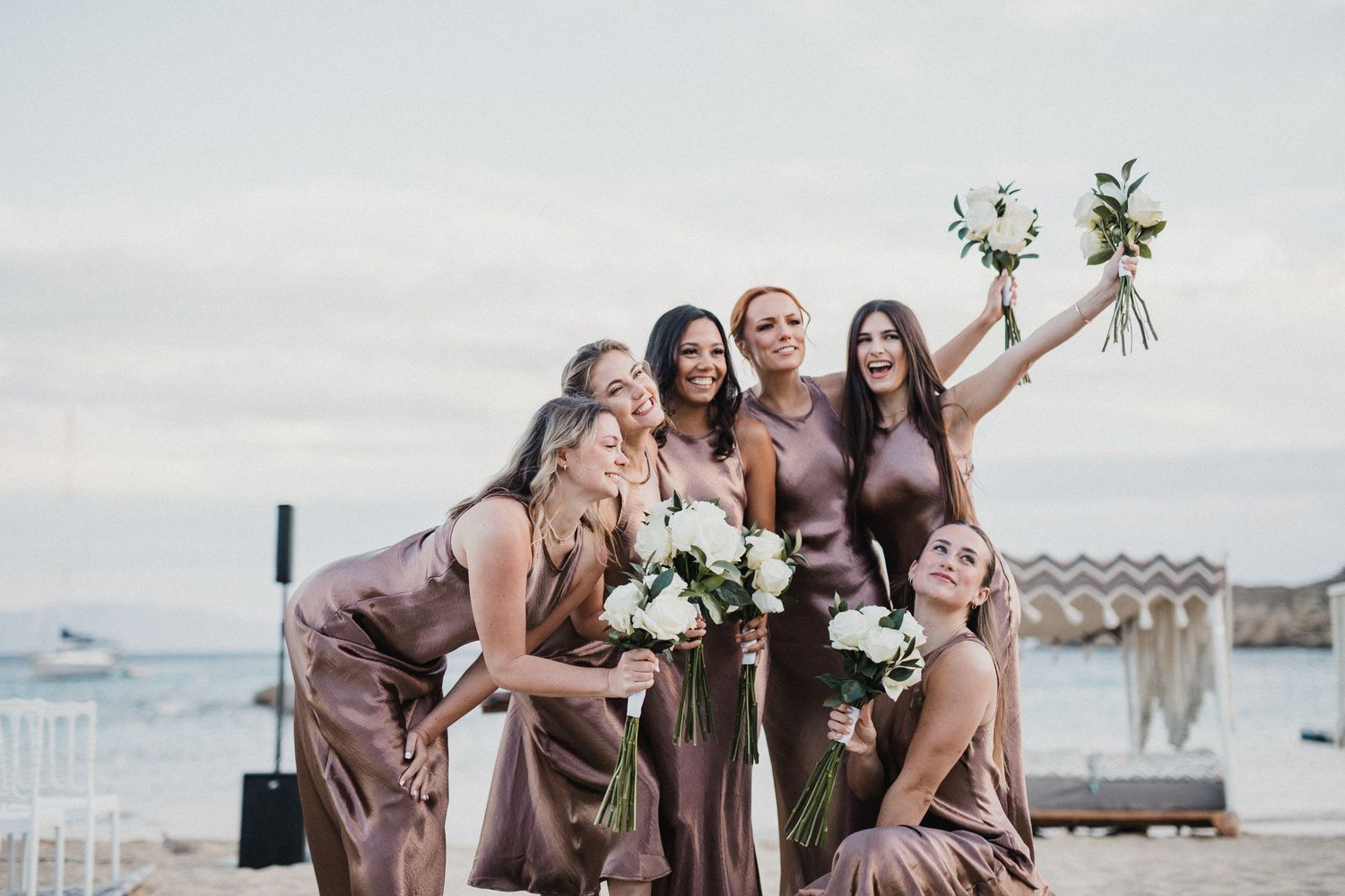 Bridesmaids in silk brown dresses posing playfully with bouquets by the ocean.