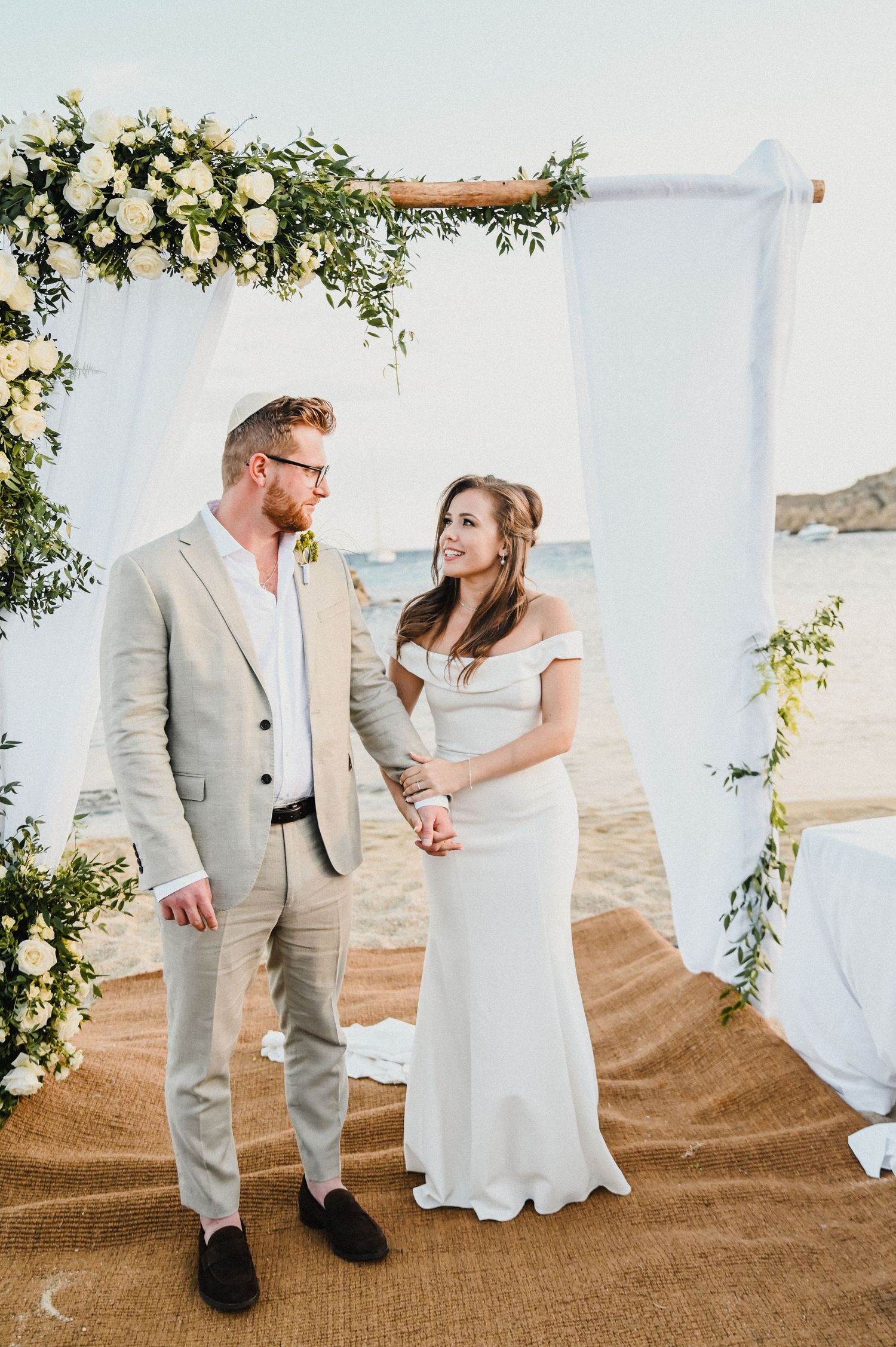 Wedding couple stands under floral arch decorated with white roses and greenery by the ocean.