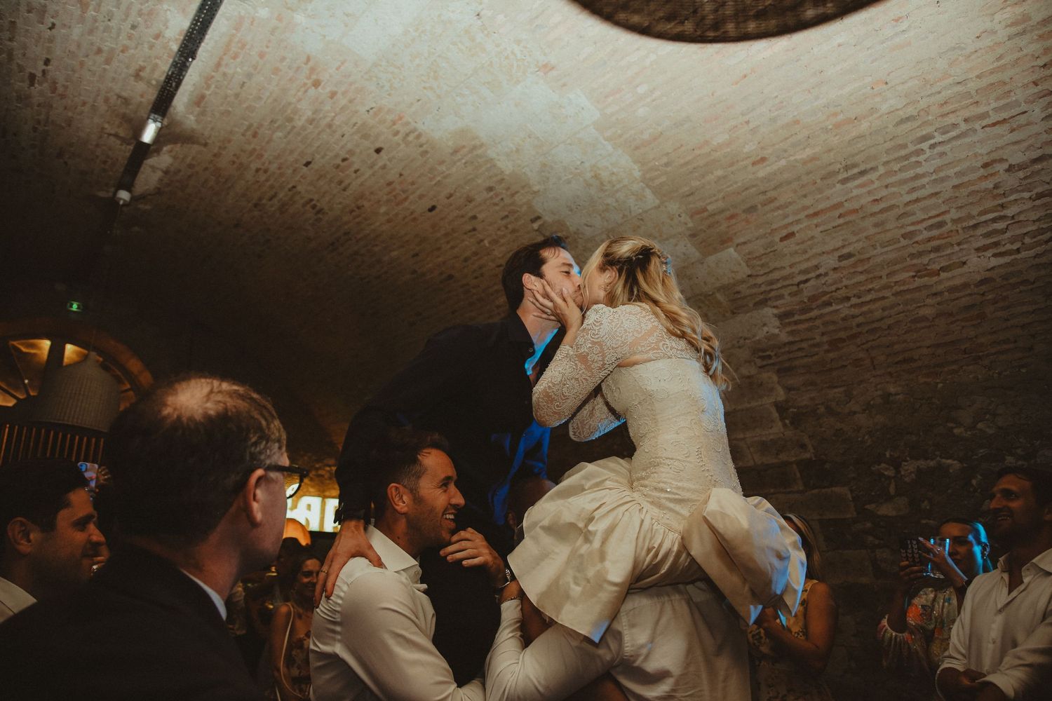 A celebratory wedding dance lift moment in a rustic underground venue with vaulted ceilings.