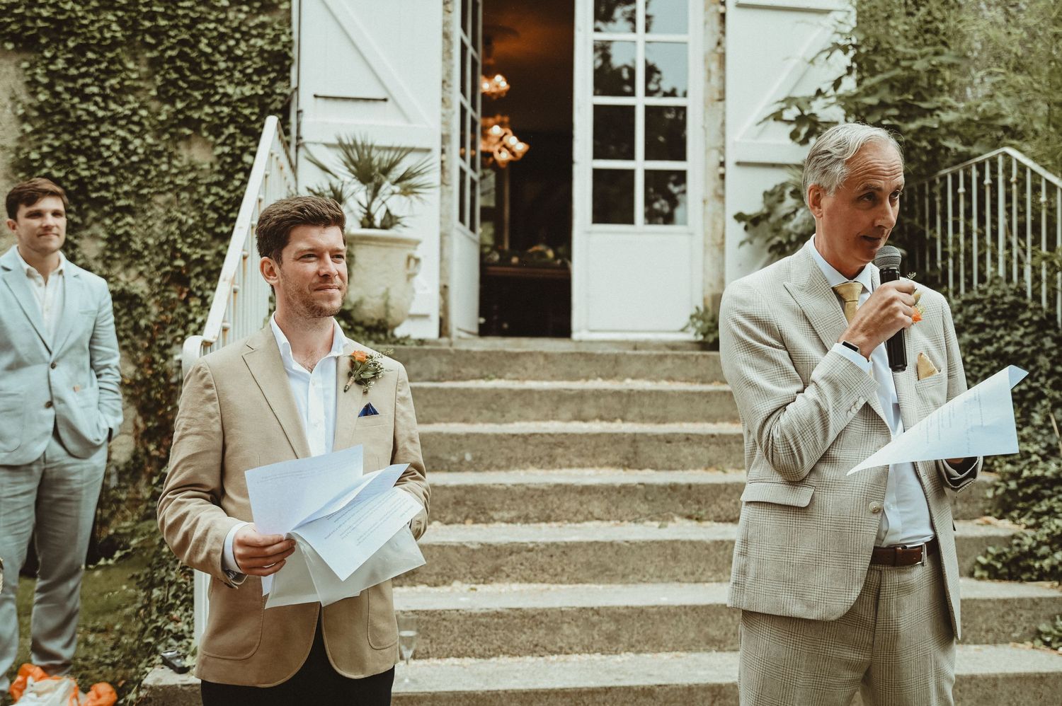 Men in light colored suits hold papers while speaking at an outdoor wedding venue.