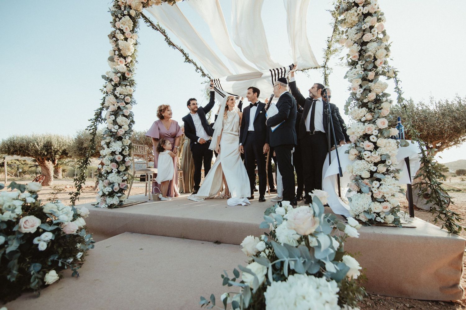 Wedding party poses under floral arch decorated with white flowers and greenery.
