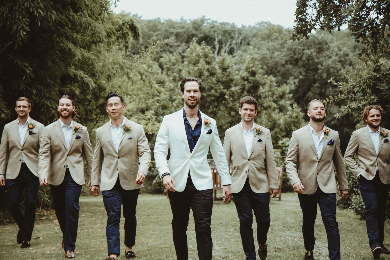 Groomsmen in beige suits walking with groom in white jacket on grassy lawn.