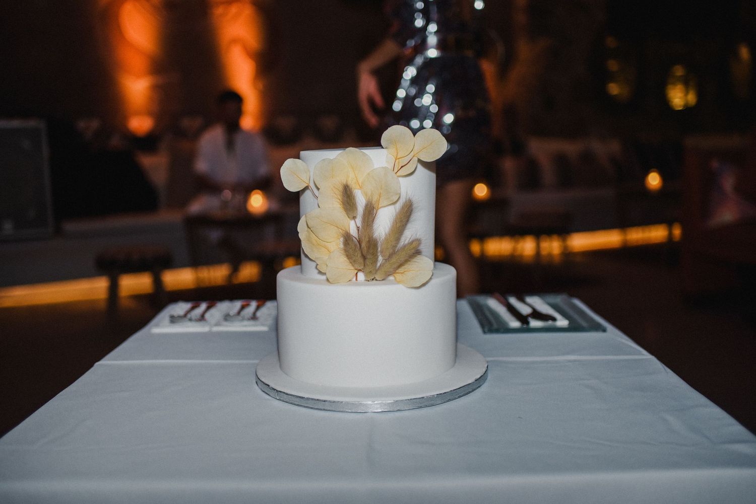Elegant white two-tier wedding cake with delicate gold leaf accents on display at an evening reception.