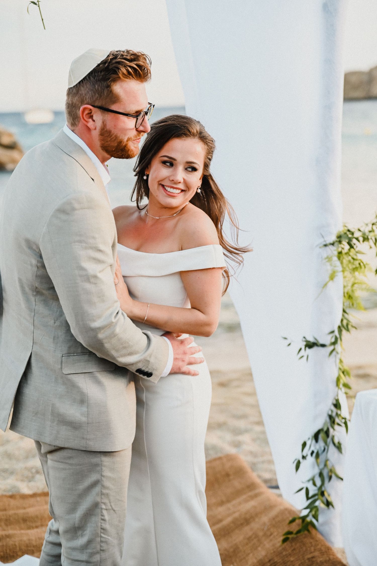 Couple embraces under floral wedding arch with seaside backdrop.
