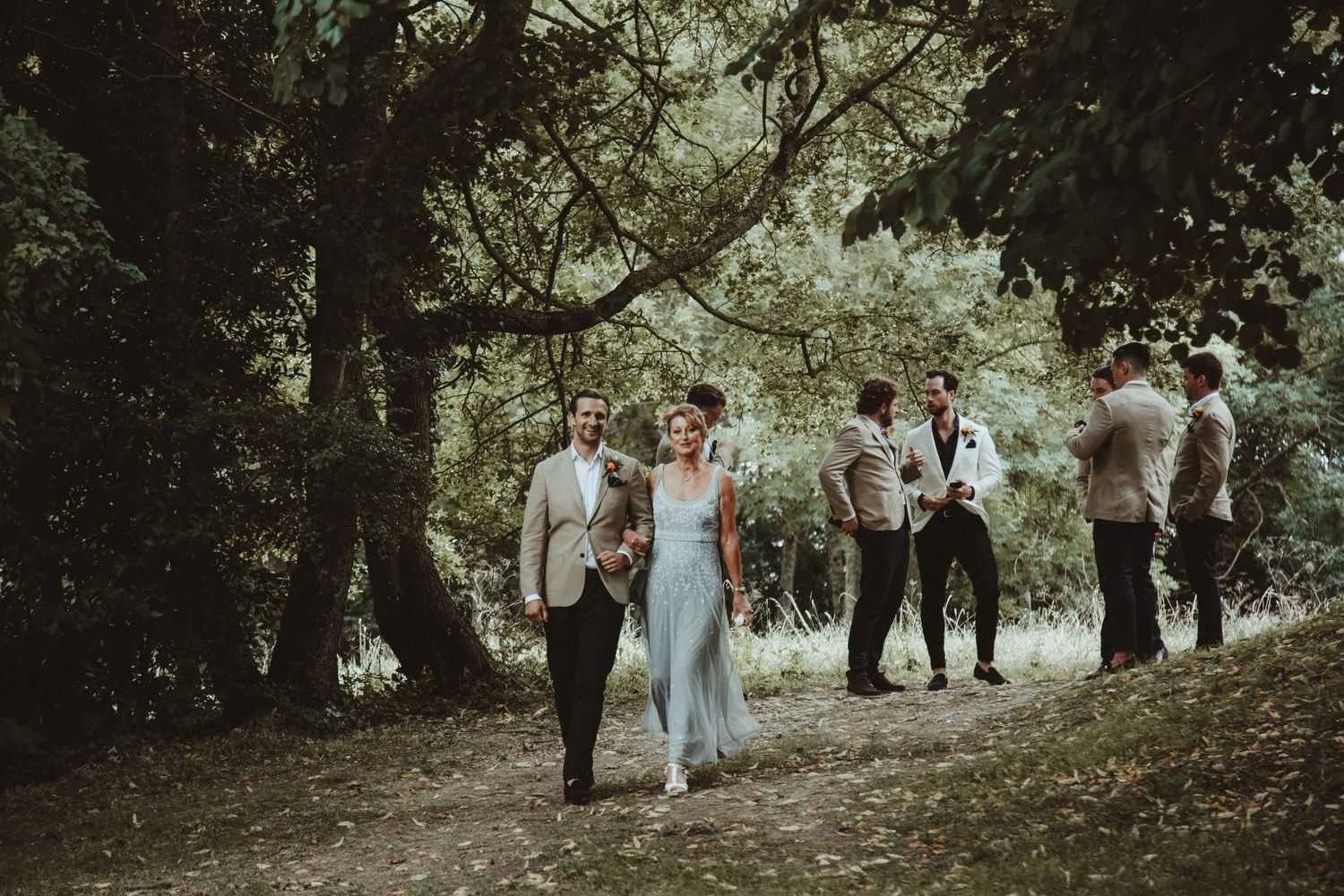 Wedding party walking casually through a tree-lined path outdoors.
