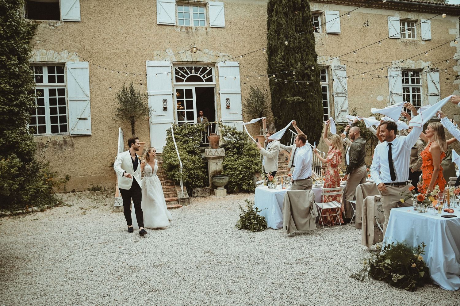 Outdoor wedding celebration with guests seated at decorated tables in front of rustic stone building.