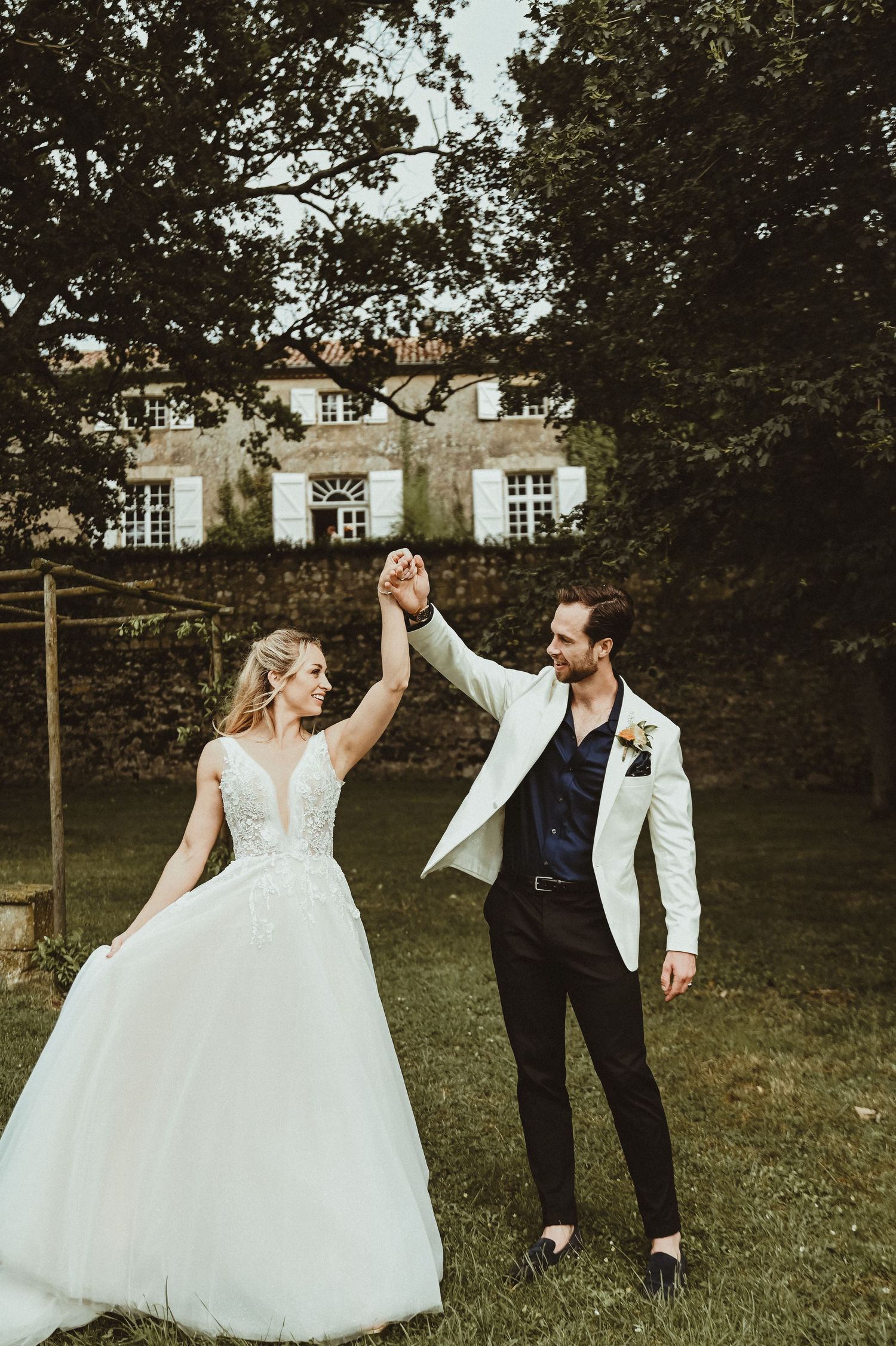 A joyful wedding couple dances on a lawn with a beautiful stone mansion in the background.