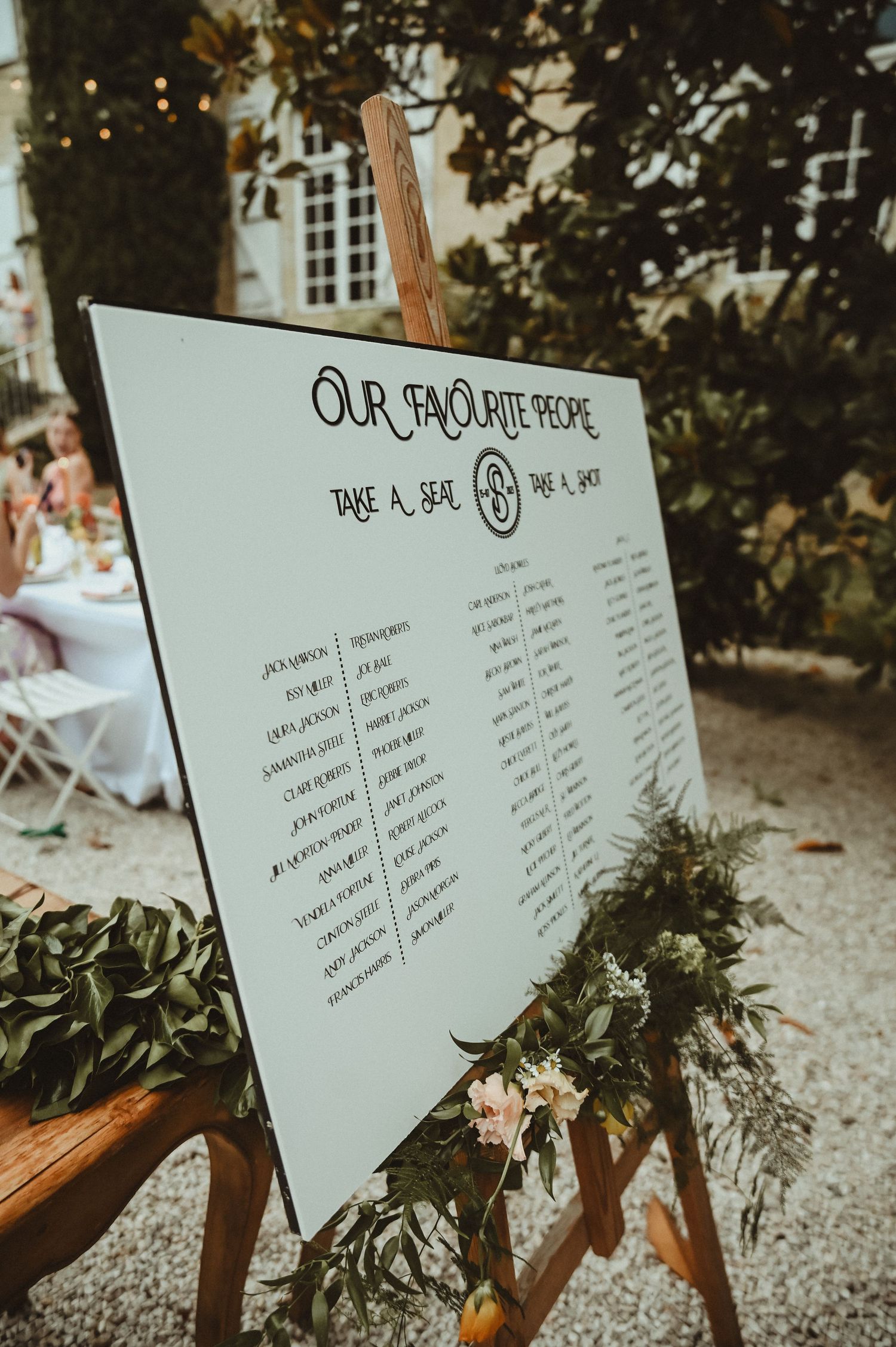 Wedding seating chart display on white board with floral decorations in rustic outdoor setting.