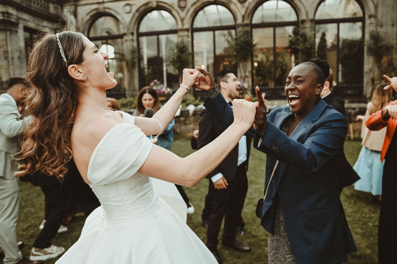 Wedding guests dance and celebrate on the lawn of an elegant stone building.