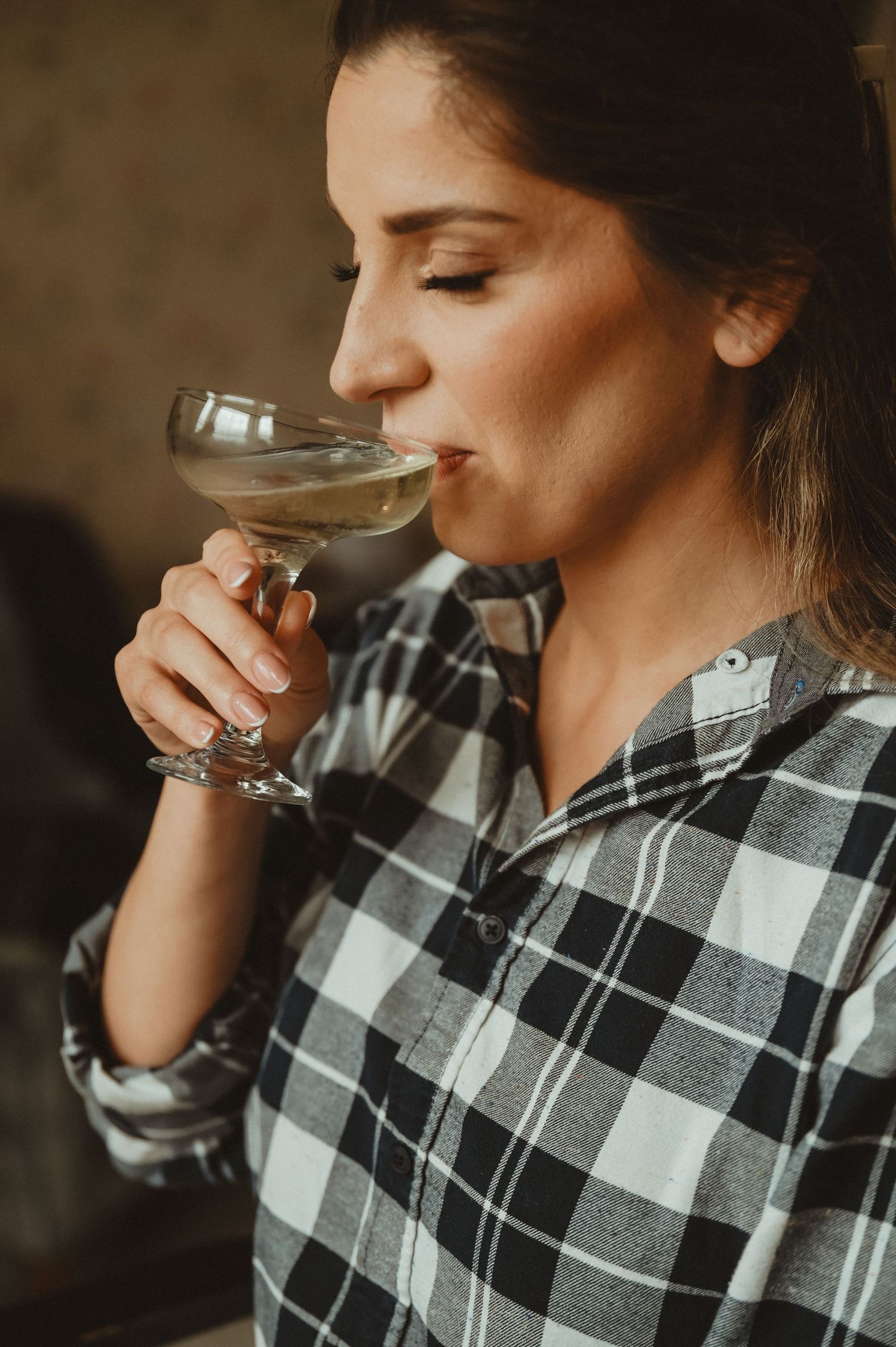 A person in a plaid shirt sips from a vintage-style cocktail coupe.
