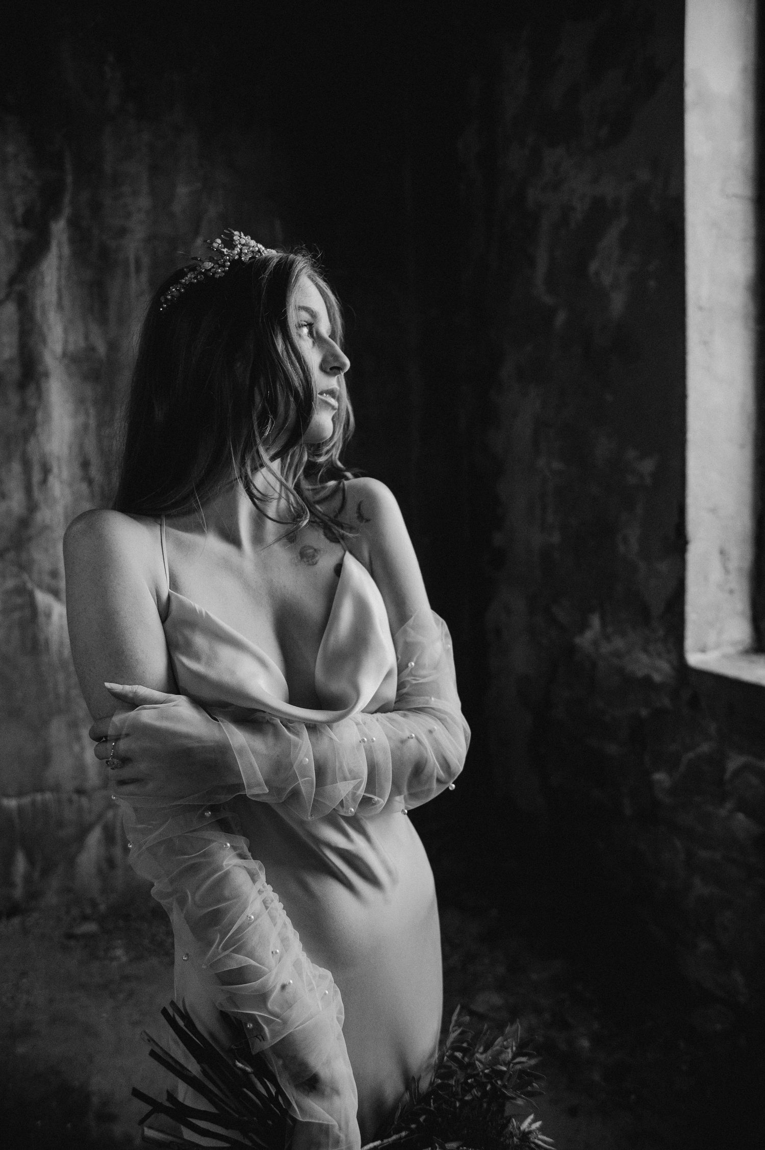Artistic monochrome photo of bride in sleek dress with sheer sleeves looking towards window.