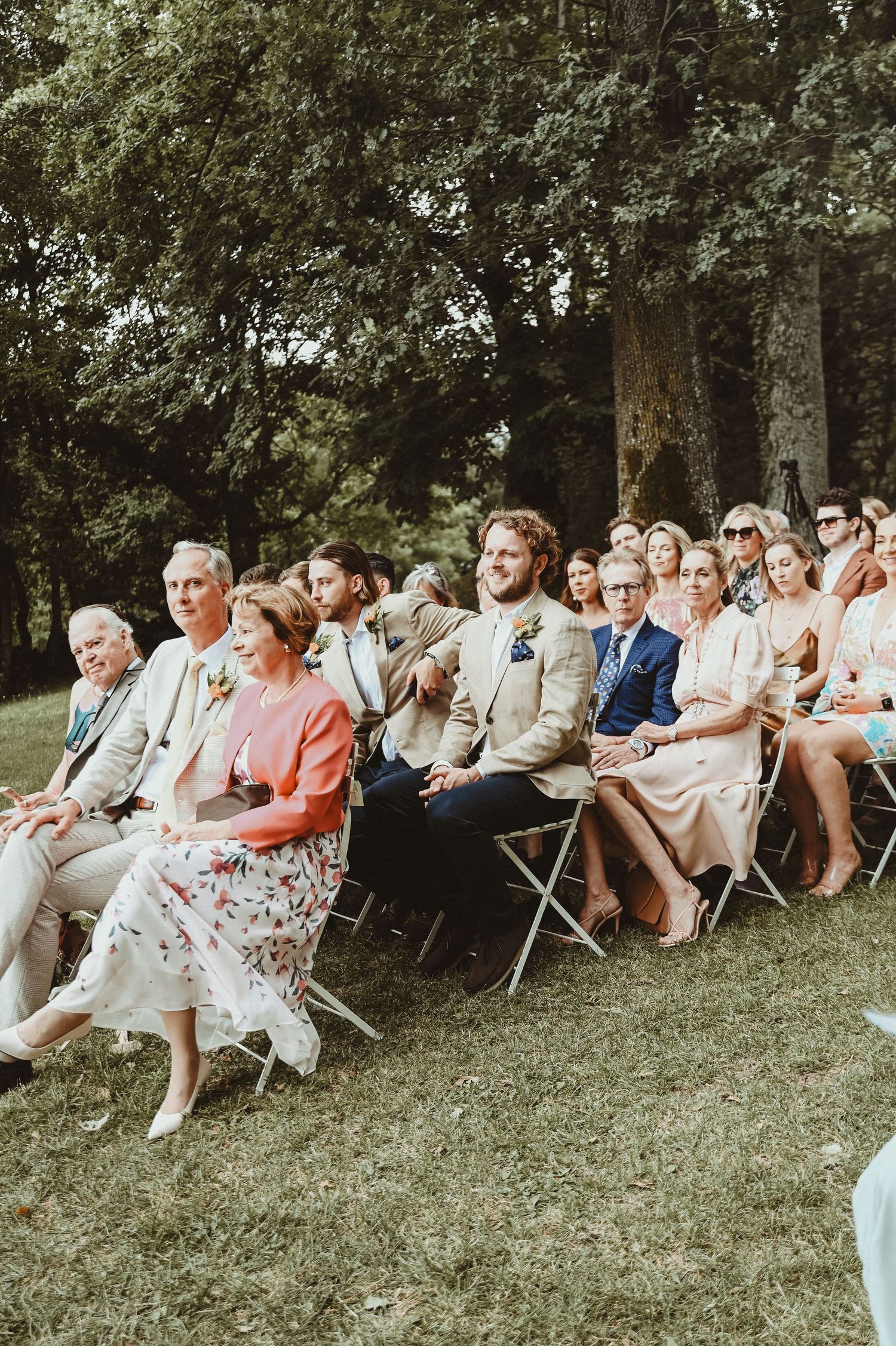 Wedding guests seated on folding chairs watch the ceremony in a garden setting beneath tall trees.