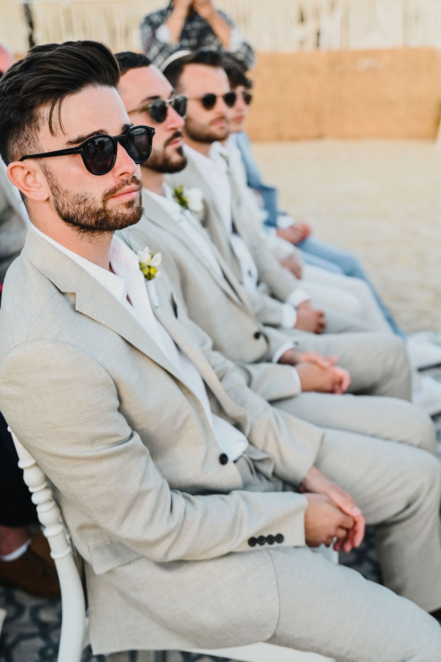 Groomsmen in light gray suits and sunglasses sit in a row at a beach wedding ceremony.