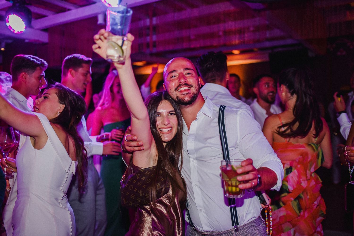 Party-goers celebrating and dancing with drinks under vibrant purple nightclub lighting.