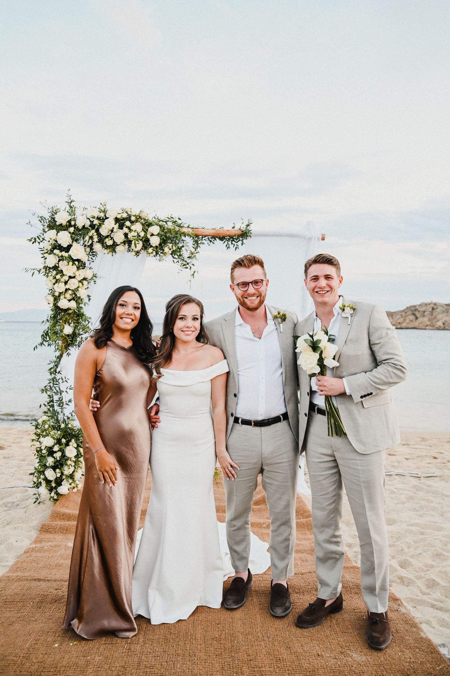 Small group poses under white floral wedding arch on beachfront platform.