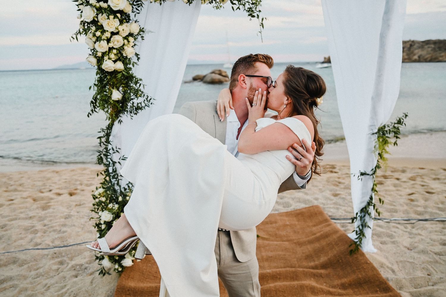 Playful wedding moment under floral arch as groom lifts bride on beach.