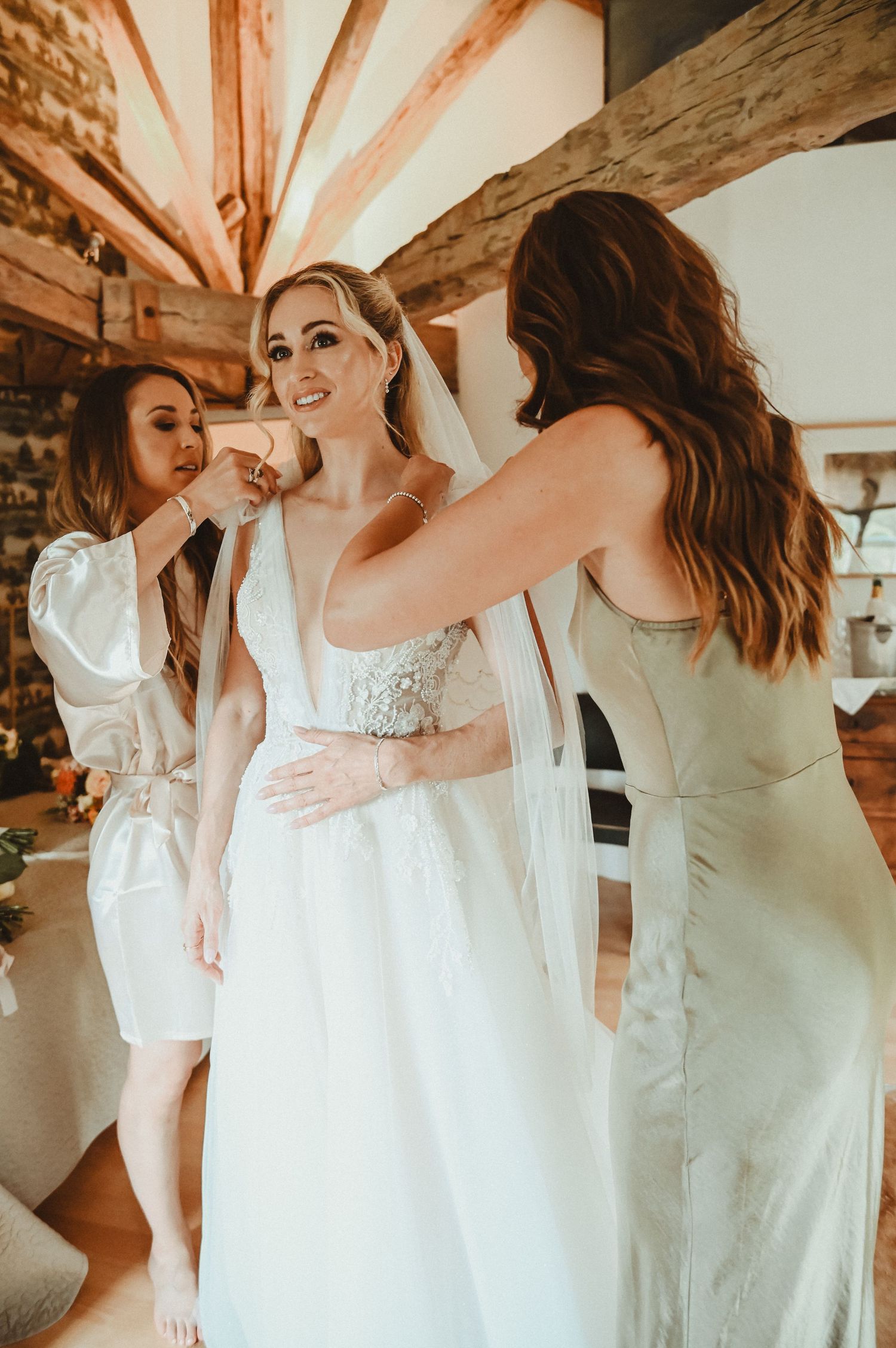 Wedding preparation scene with attendants helping adjust a white lace gown in a rustic room with exposed beams.