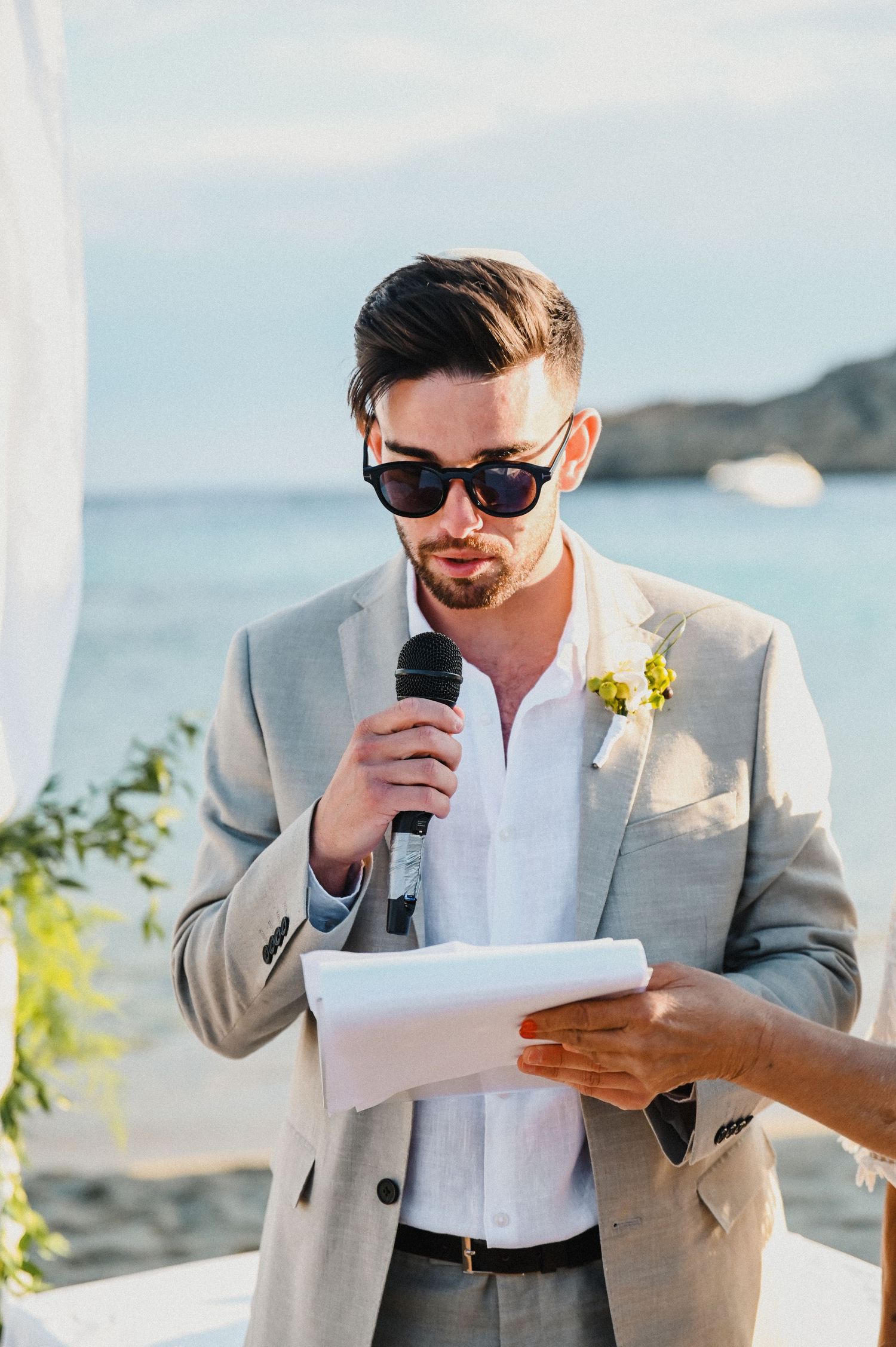 Wedding speaker in grey suit and sunglasses delivers speech at outdoor beach ceremony.