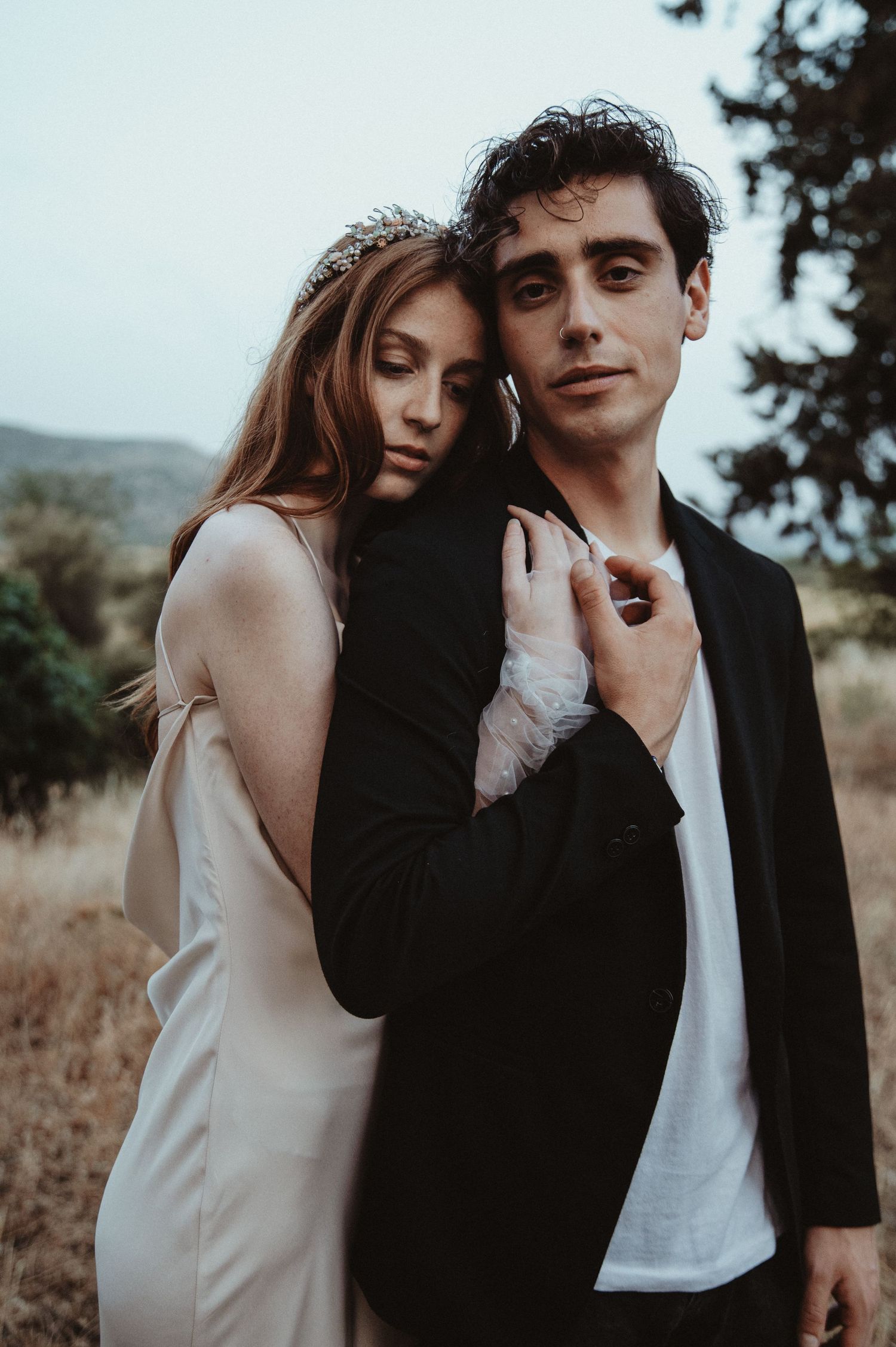 A romantic couple poses together outdoors in formal attire with moody lighting and natural scenery.