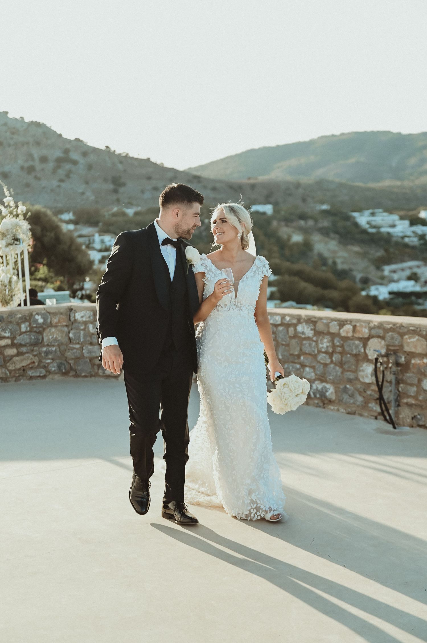 Elegant couple in wedding attire walk along stone terrace with mountain views.