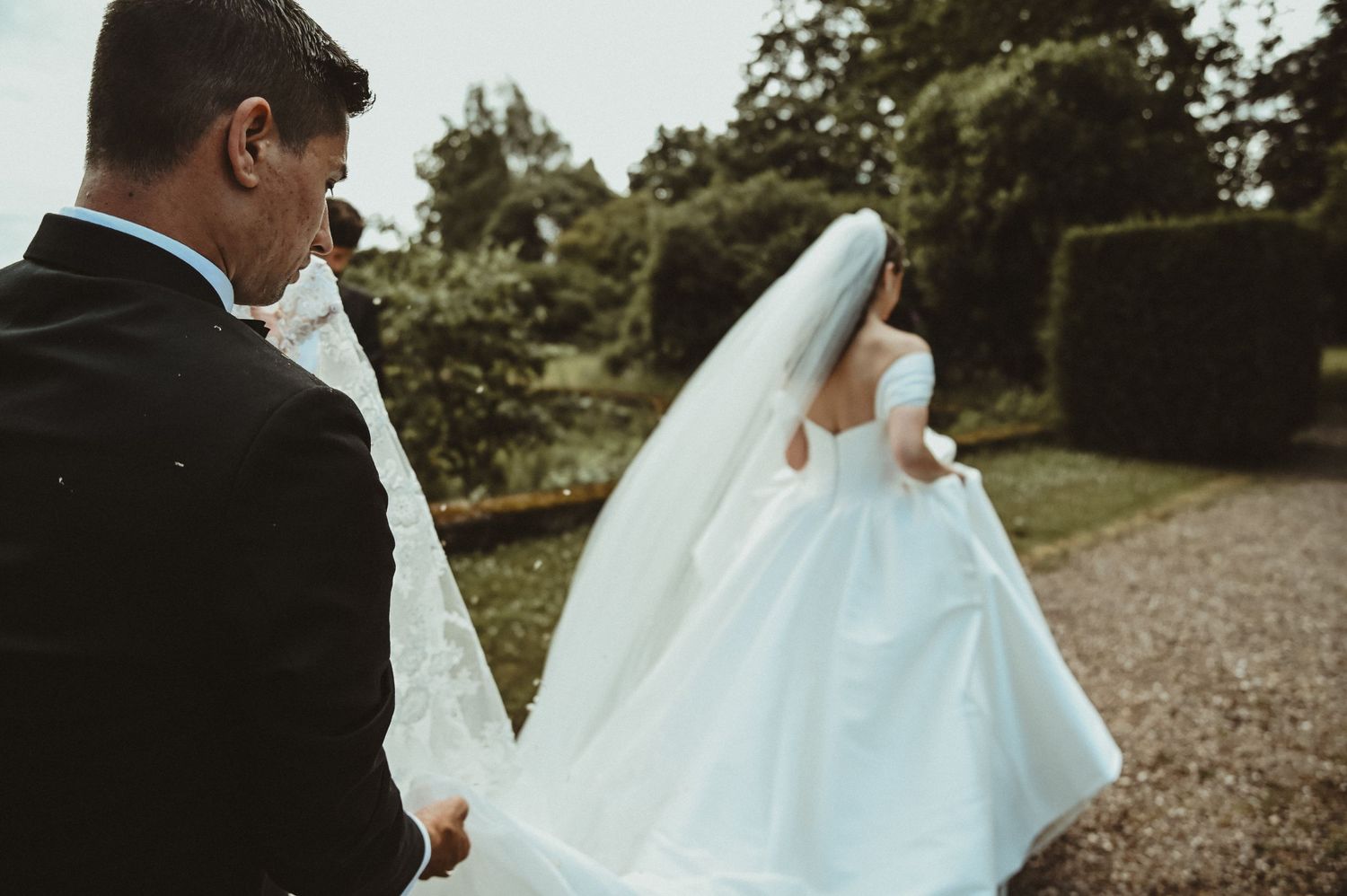 A wedding couple walks along a garden path captured from behind.