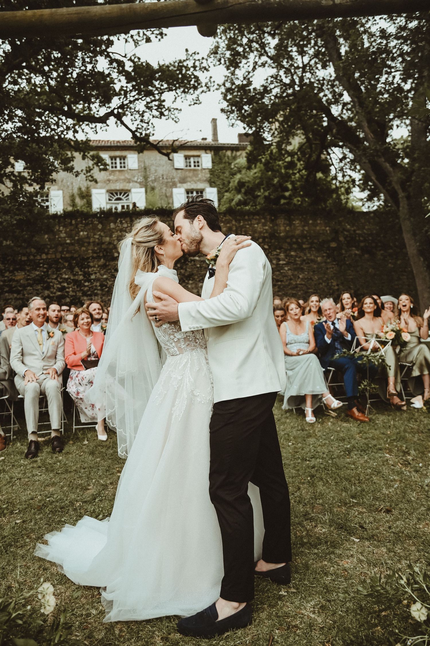 Newlyweds share their first kiss as guests look on during an outdoor garden wedding ceremony.