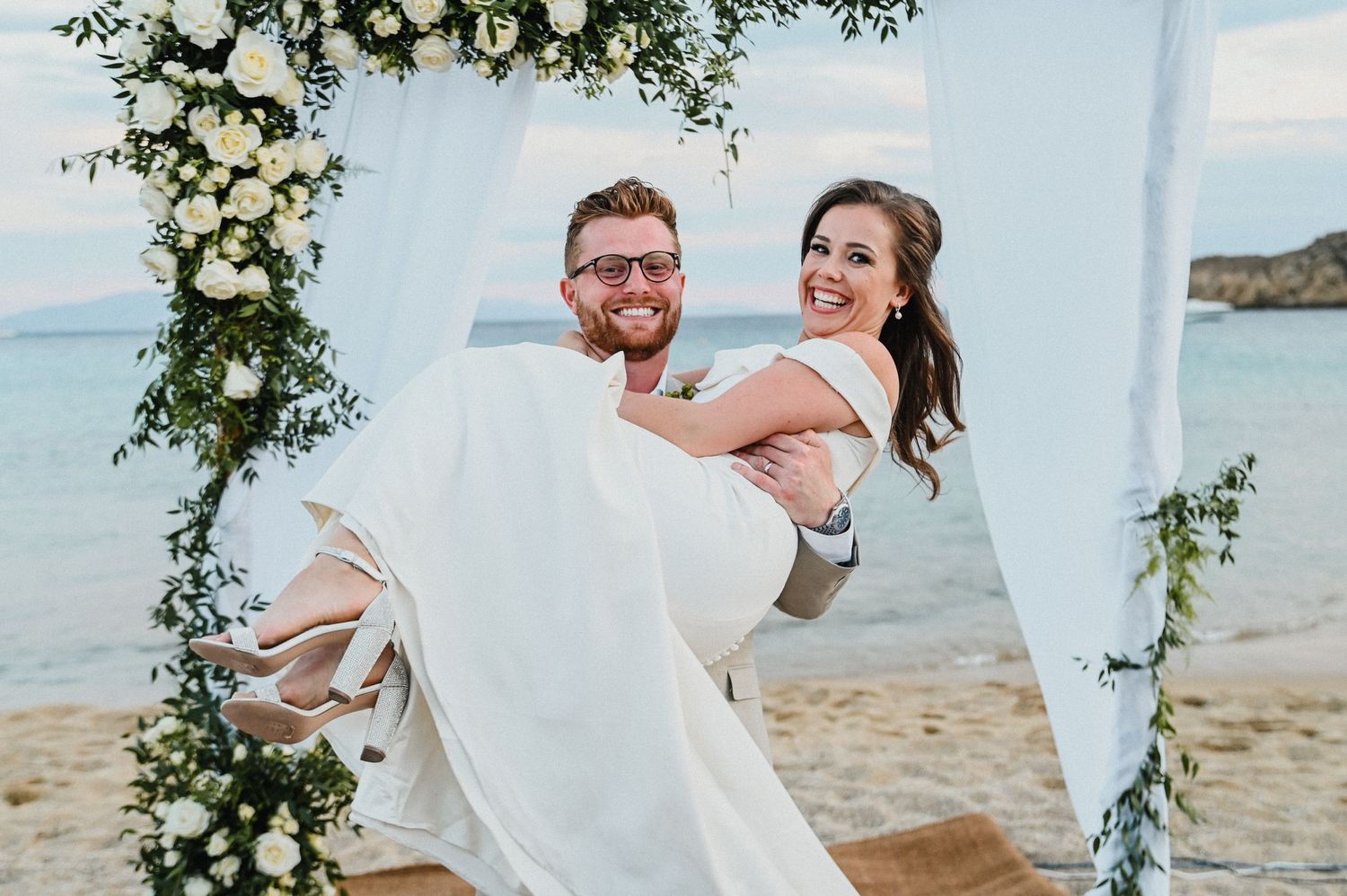 A groom carries his bride under a floral arch on a scenic beach during their wedding ceremony.